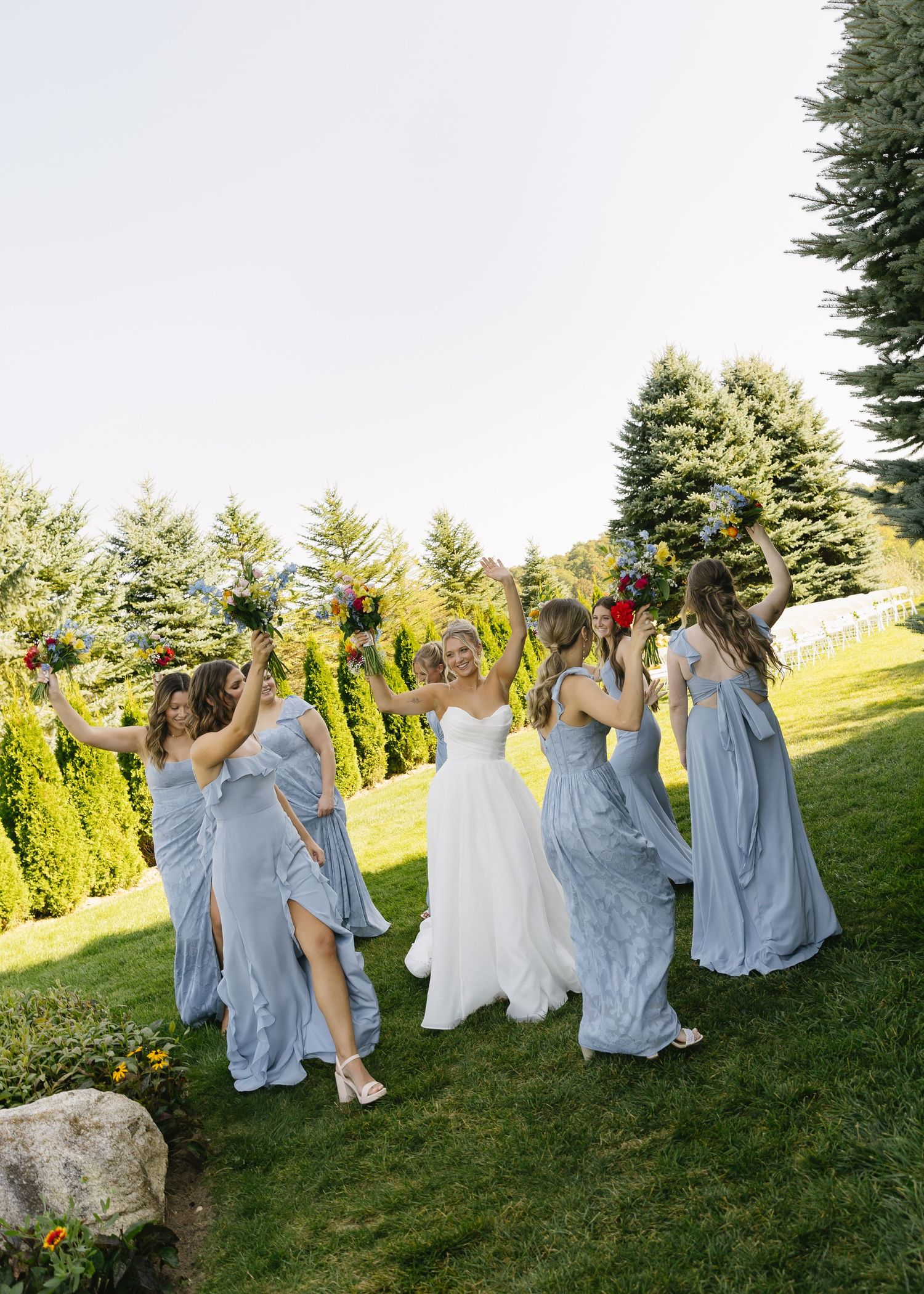 Bridal party in flowing blue dresses dancing on a green lawn.