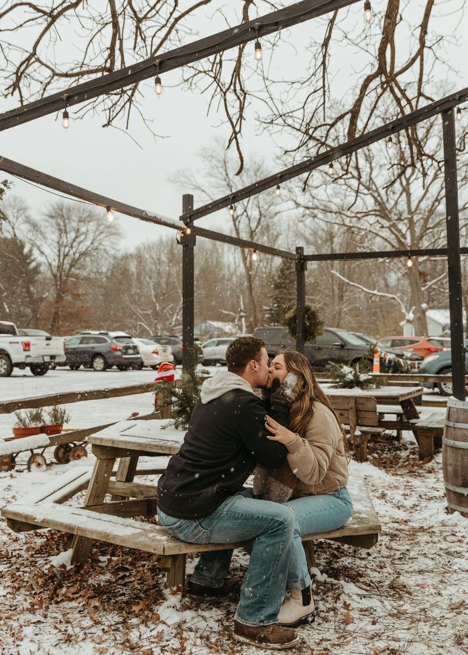 A couple embraces on wooden picnic tables surrounded by snow and bare winter trees.