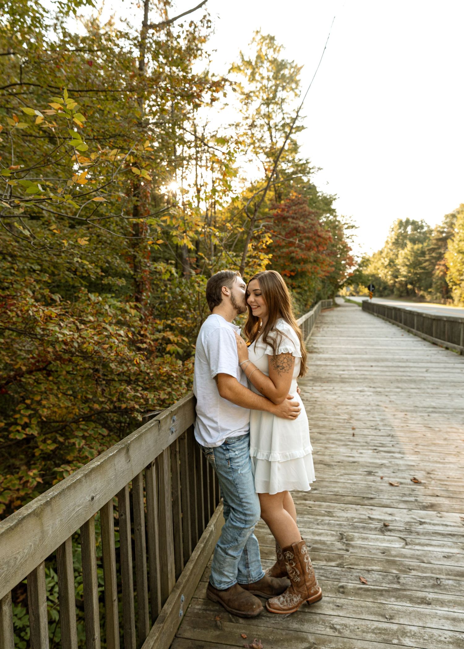A romantic couple embraces on a wooden boardwalk surrounded by autumn foliage during sunset.