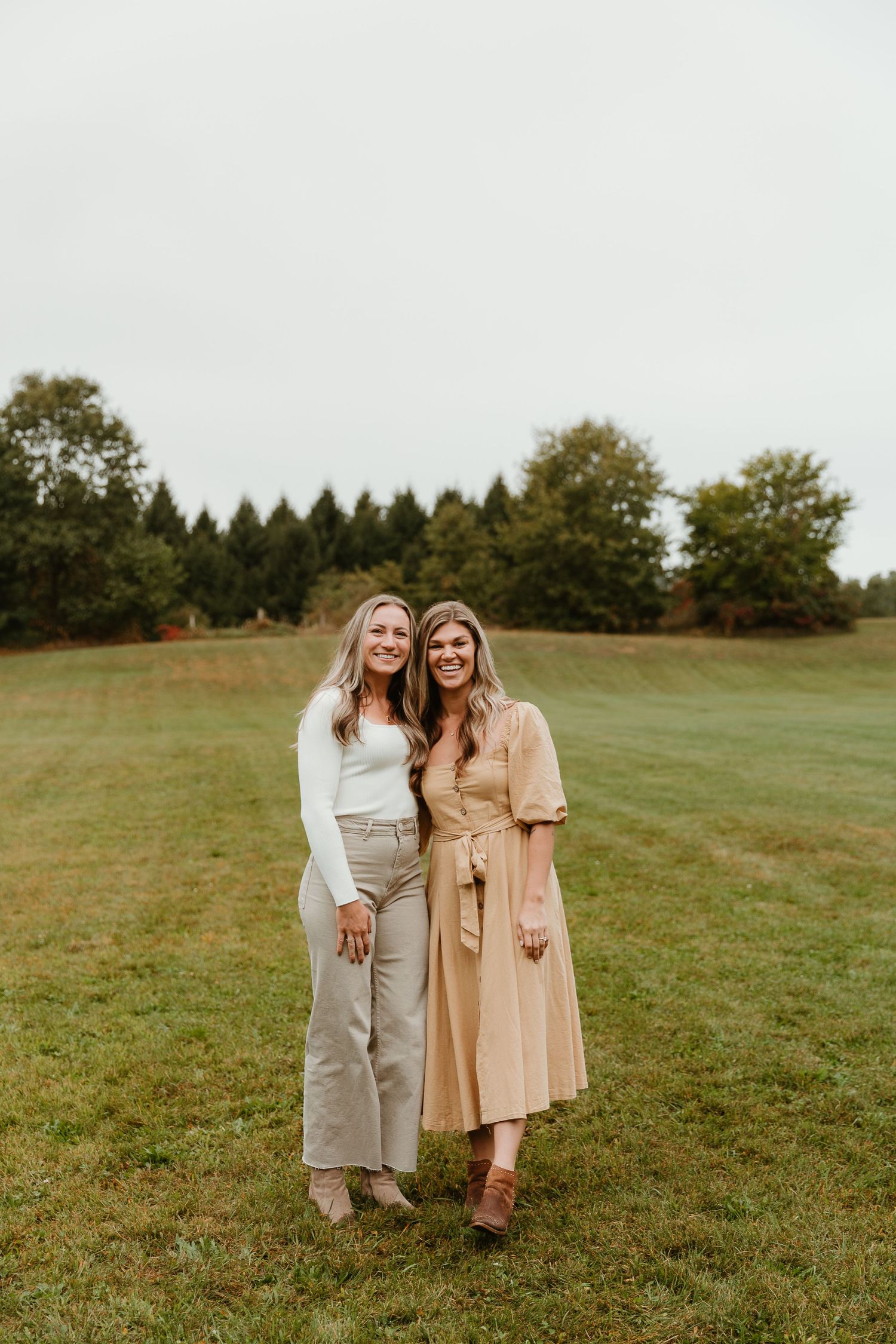 Two people in casual fall outfits embrace while standing in a grassy field on an overcast day.