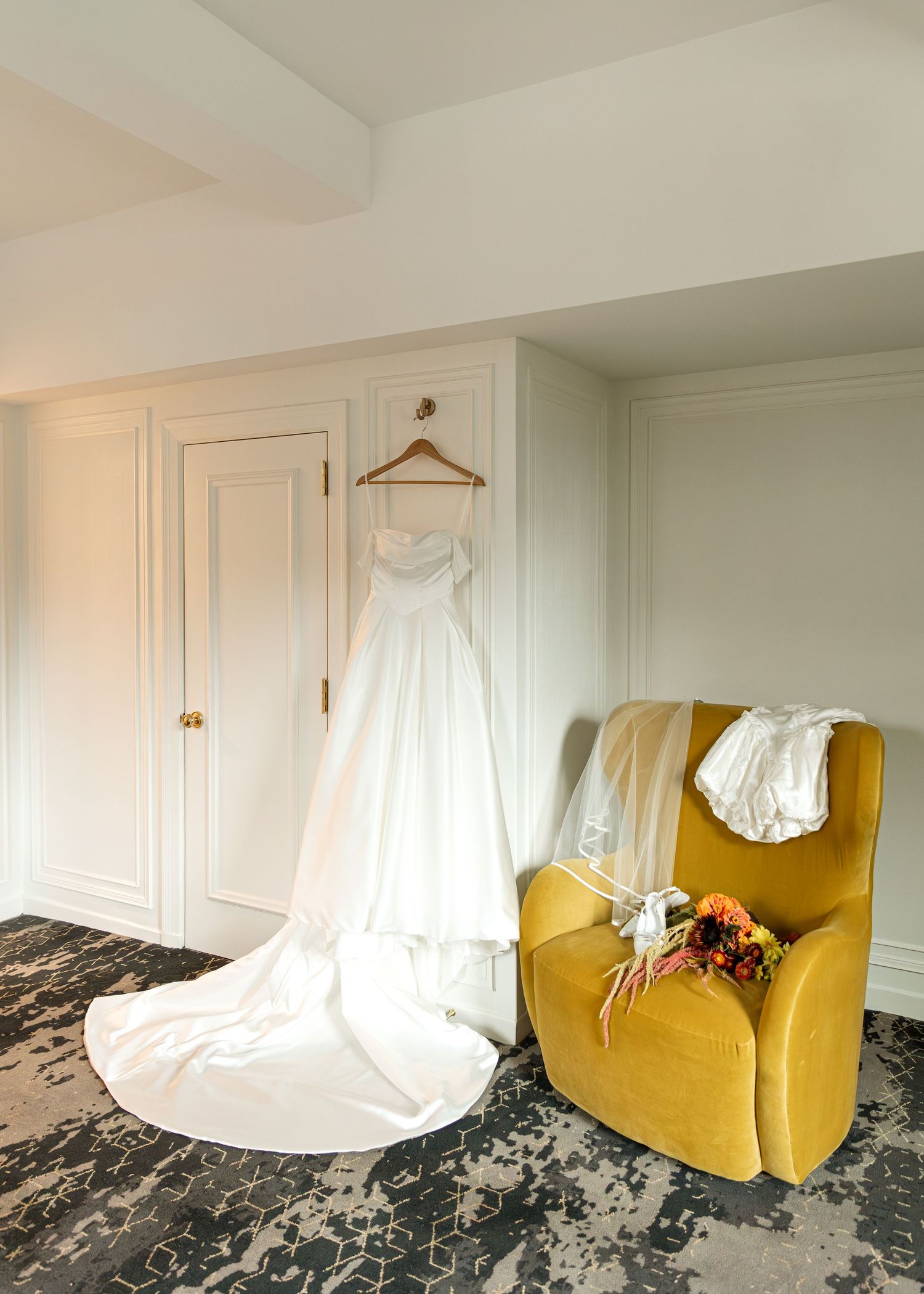 White wedding dress hangs elegantly near a yellow accent chair in a bright room.
