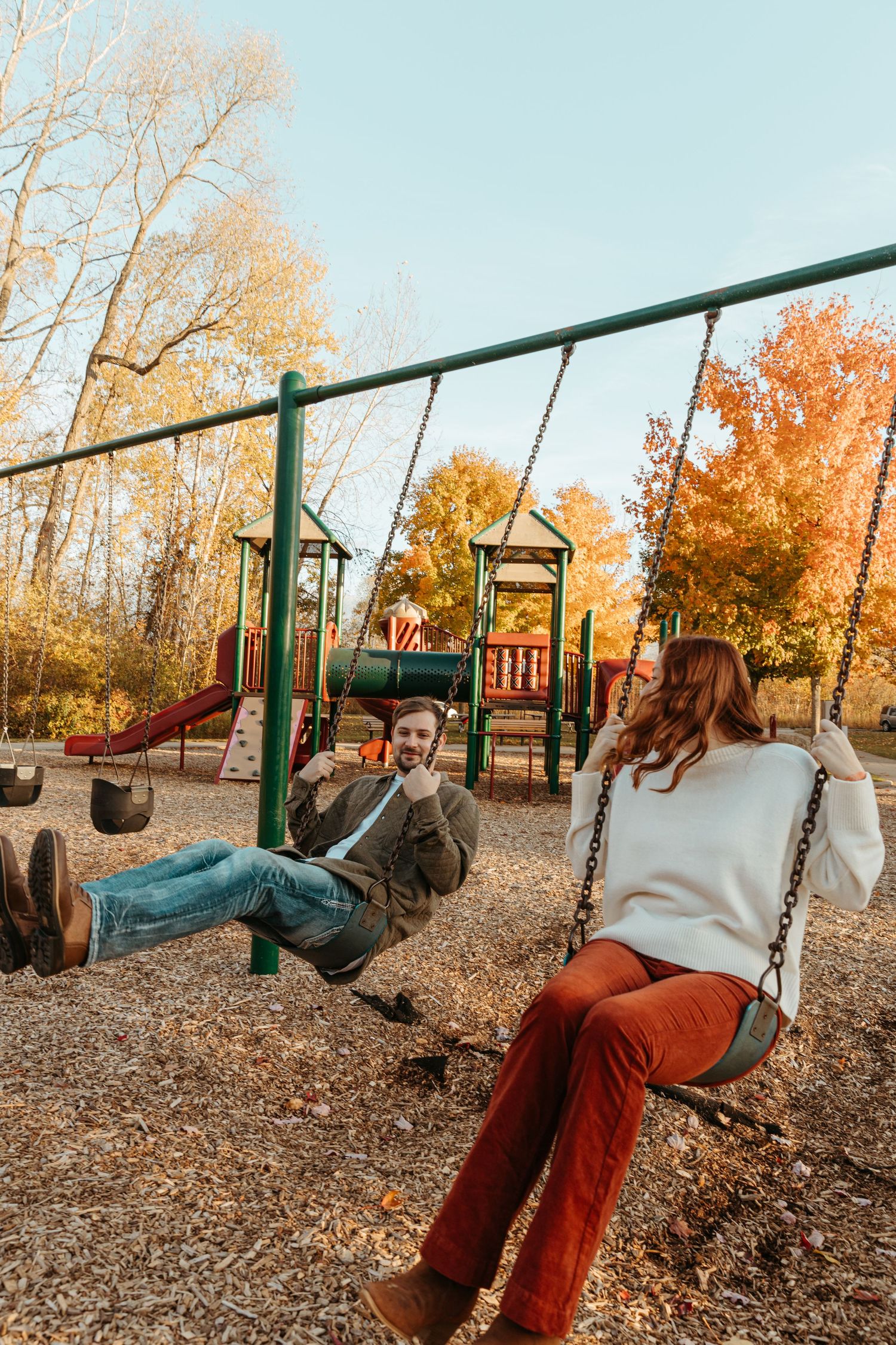 Two people swing together at a playground surrounded by fall foliage.