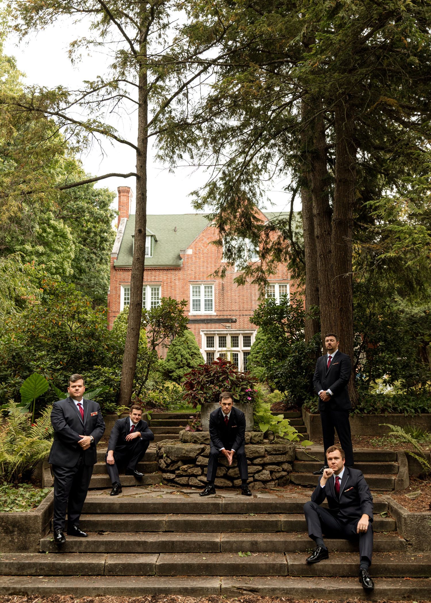 Wedding party members pose casually on outdoor stone steps surrounded by trees.