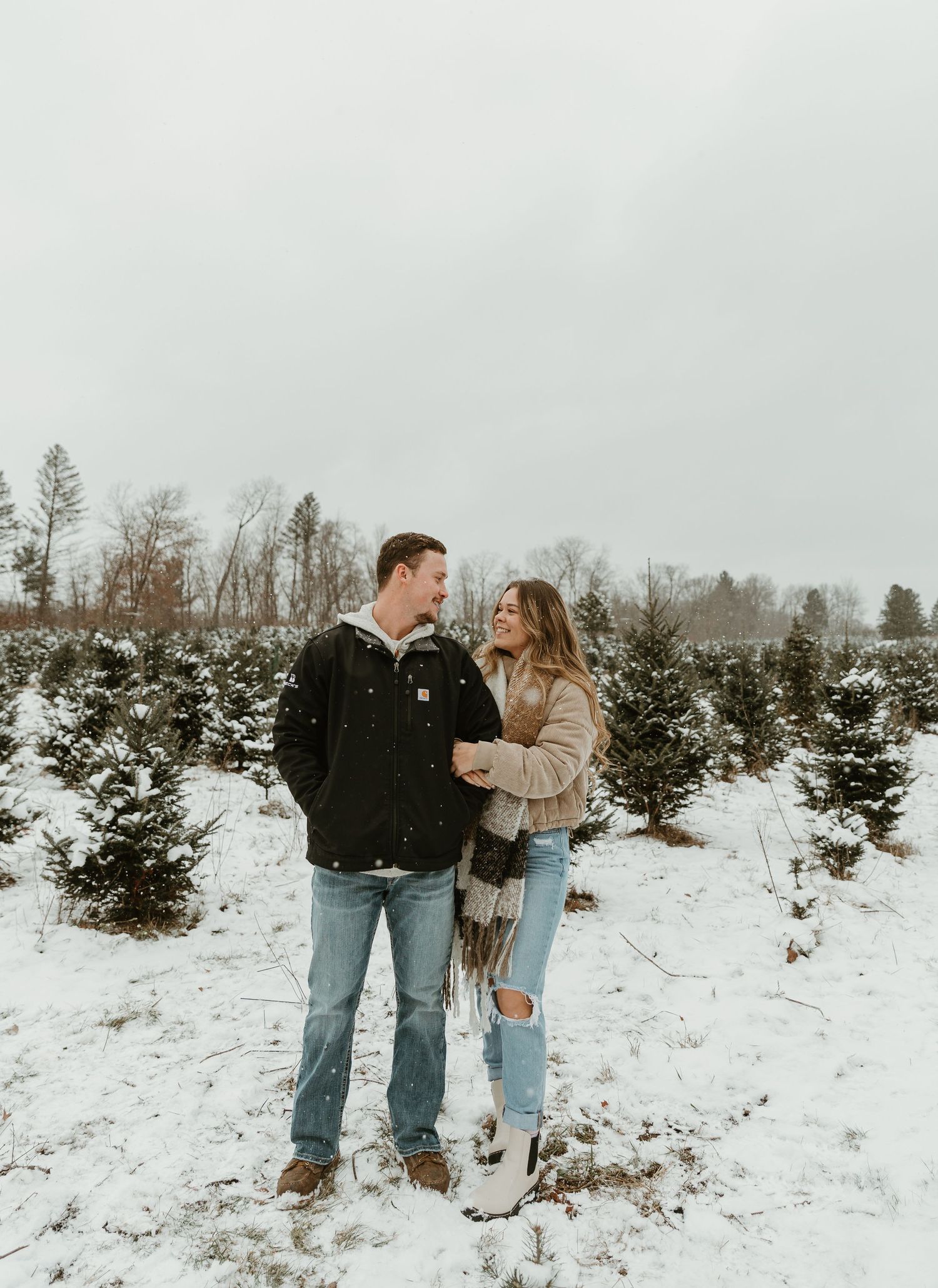 Couple sharing intimate moment in snowy Christmas tree farm during winter photoshoot.