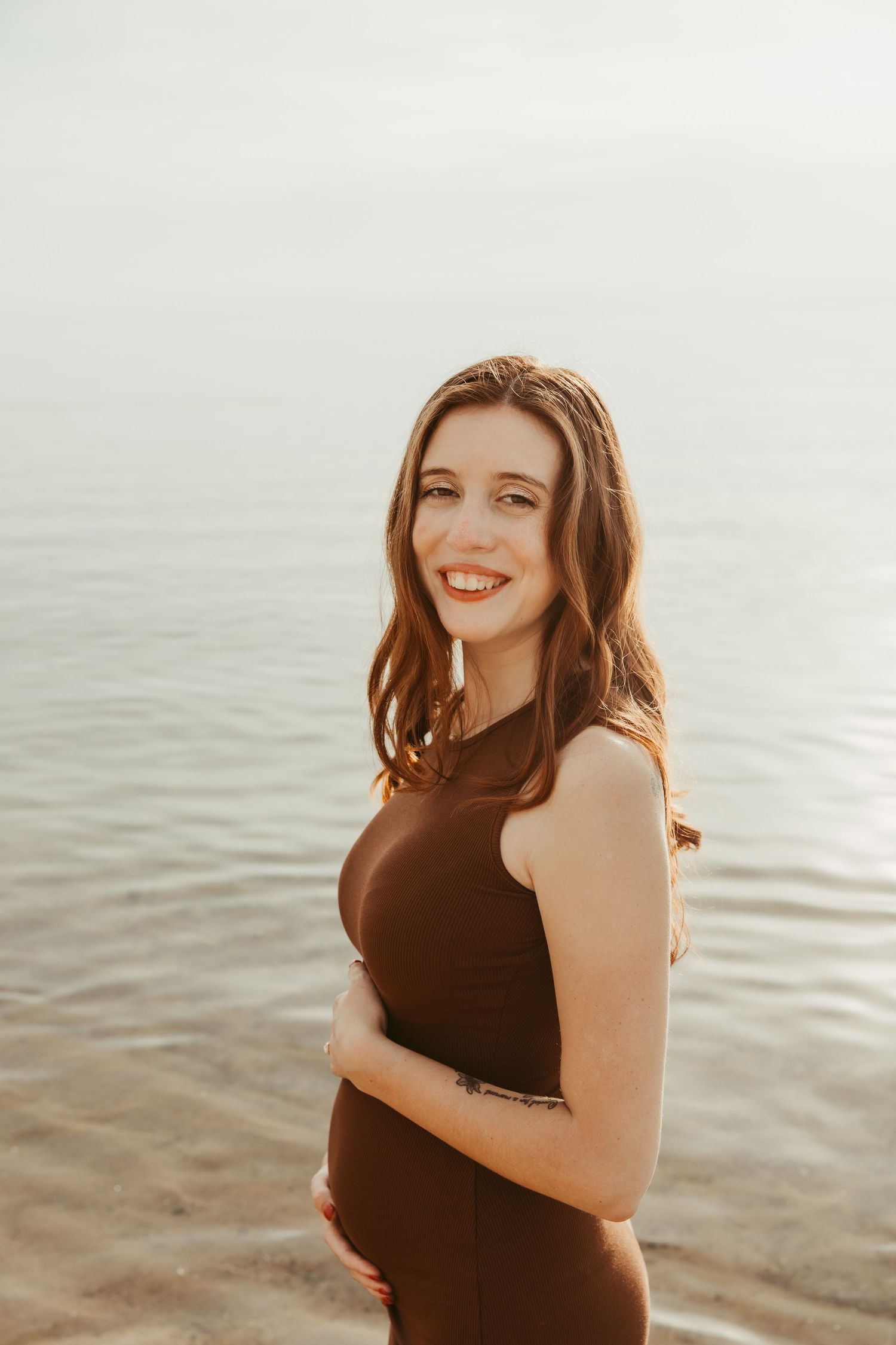Expectant mother in black dress smiling joyfully by the ocean at sunset.