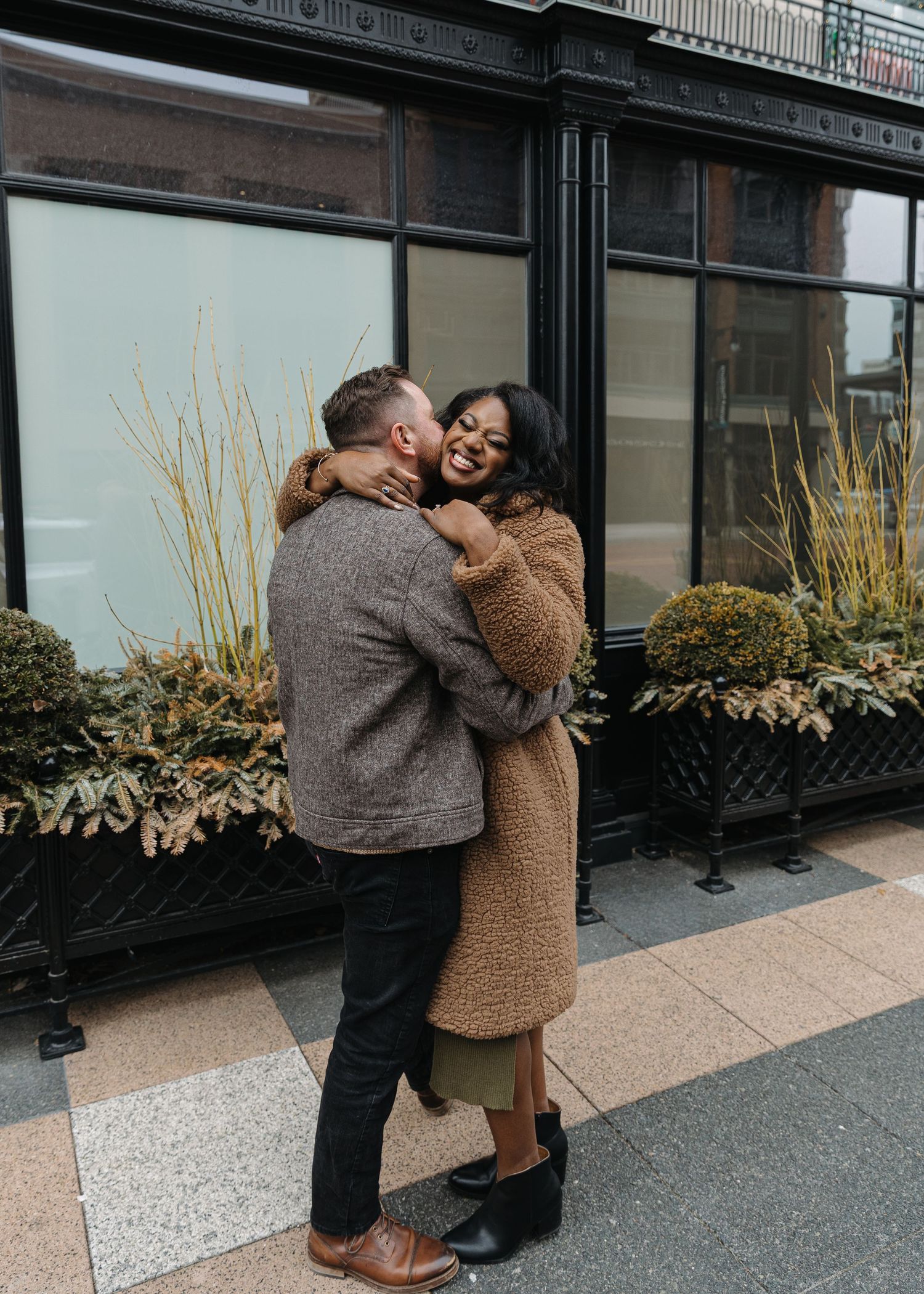 A joyful couple embraces and laughs outside a building with large black windows and green planters on a cool day.