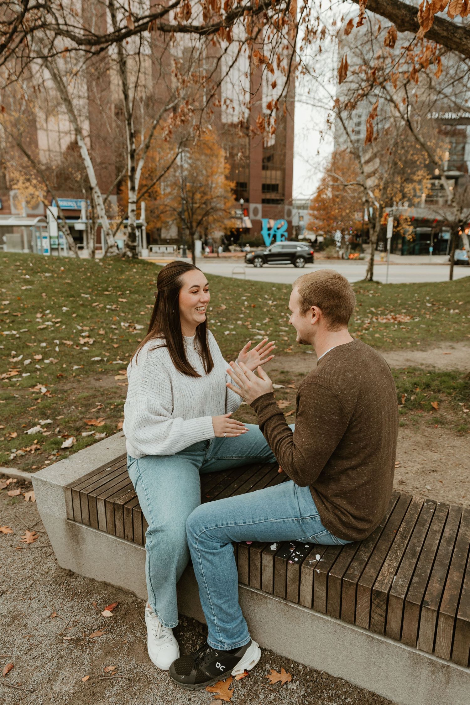 Couple sitting on a concrete bench surrounded by fall trees and fallen leaves.