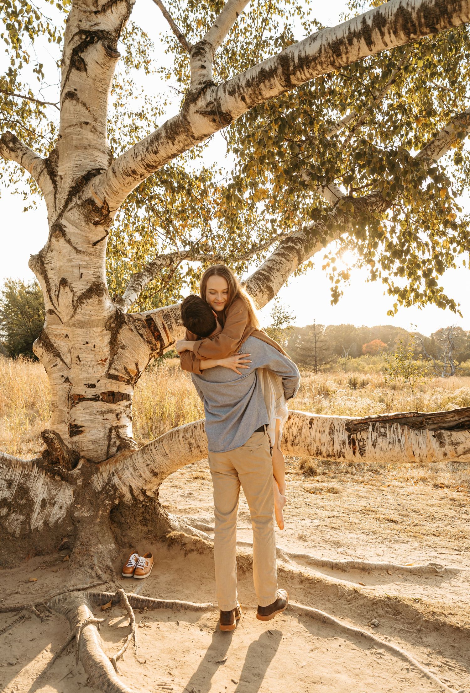 Couple embracing by a large birch tree during golden hour with their dog nearby.