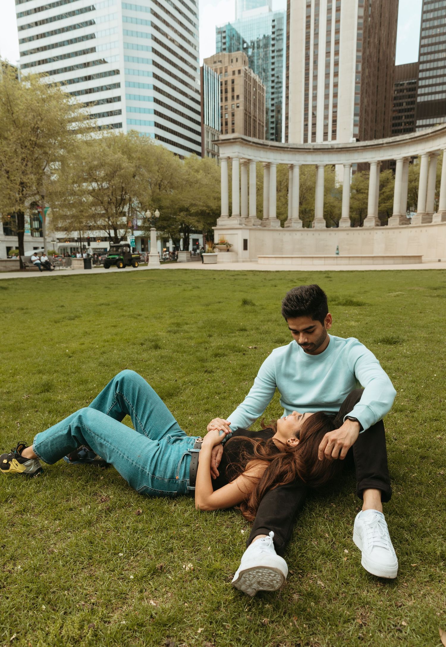 Couple lying on grass with urban skyline and buildings in background during casual photoshoot.