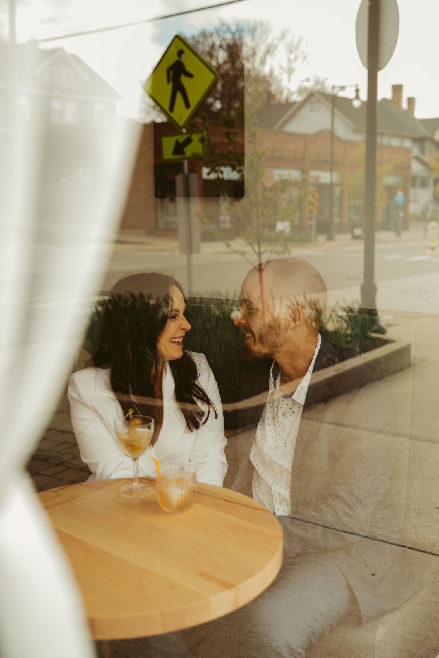 A couple shares joyful moments at a cafe table seen through a window reflection.