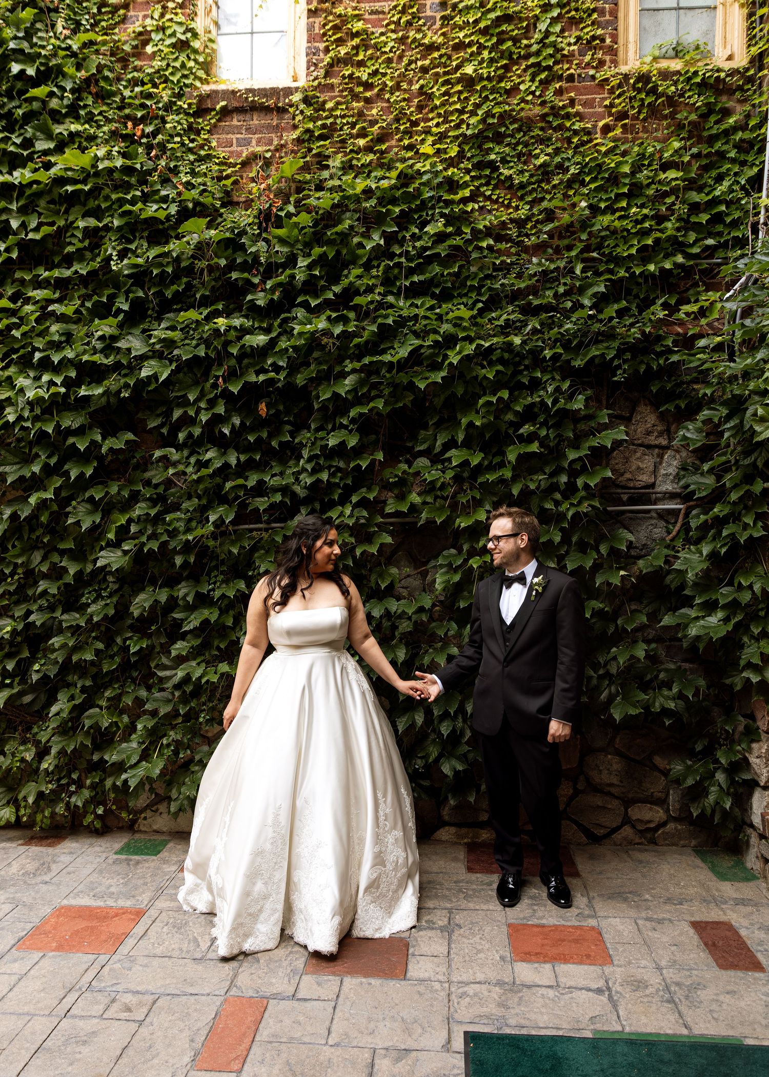 A wedding couple poses against an ivy-covered wall on a stone patio.
