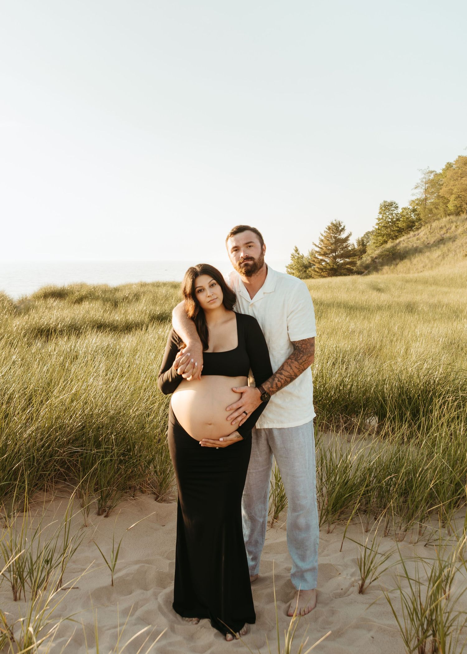 An expectant couple shares an intimate moment on coastal dunes during sunset.