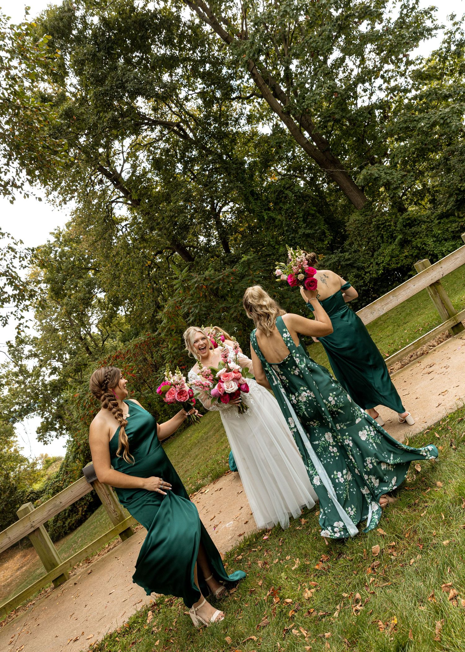 Bridesmaids in emerald green dresses pose playfully on a grassy slope during an outdoor photo session.