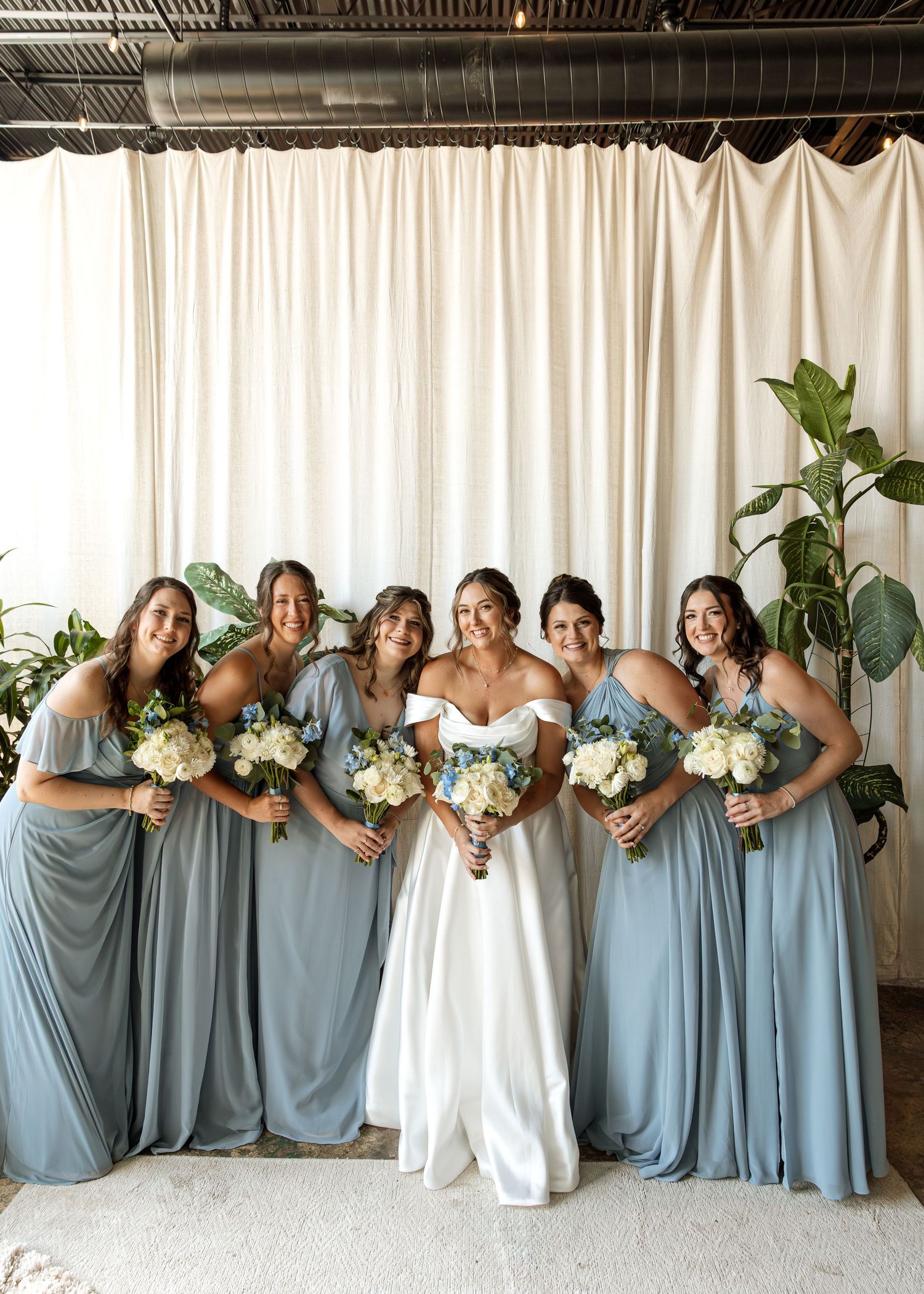 Bridal party in flowing blue dresses shares joyful moment together against white backdrop.