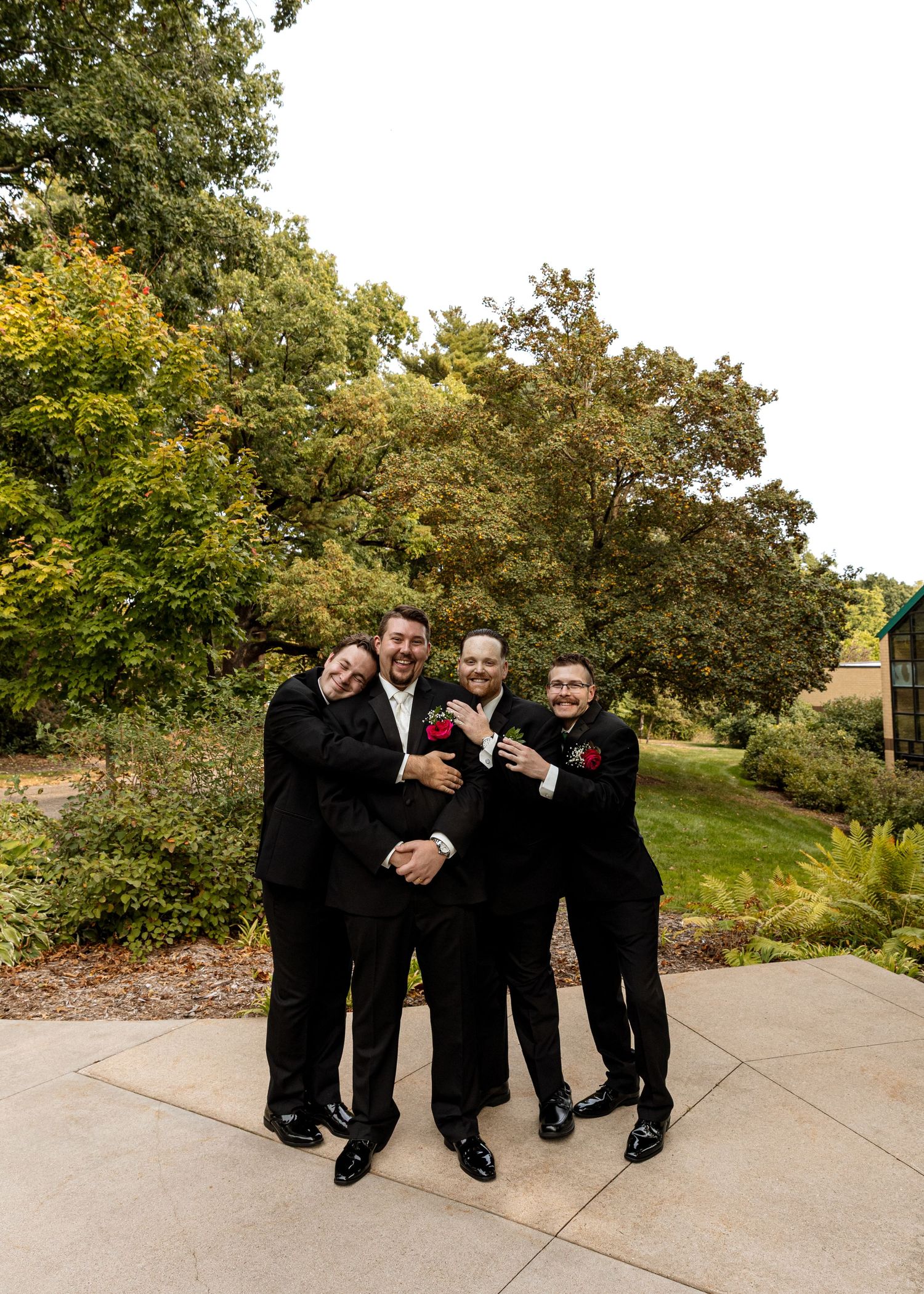 Wedding party members in black suits pose casually on concrete path with trees in background.