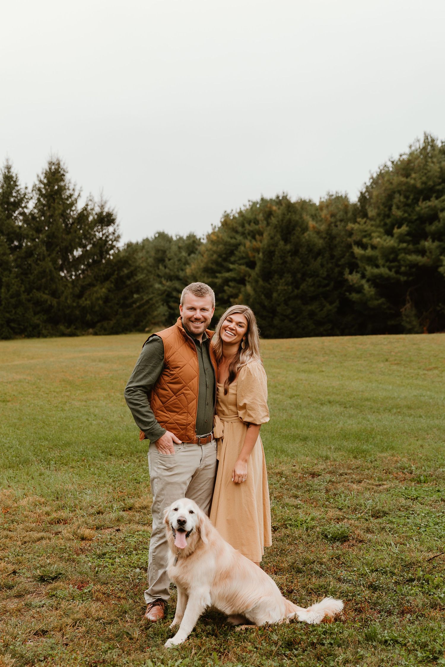 A golden retriever lies in the grass while people stand behind it in a scenic field.