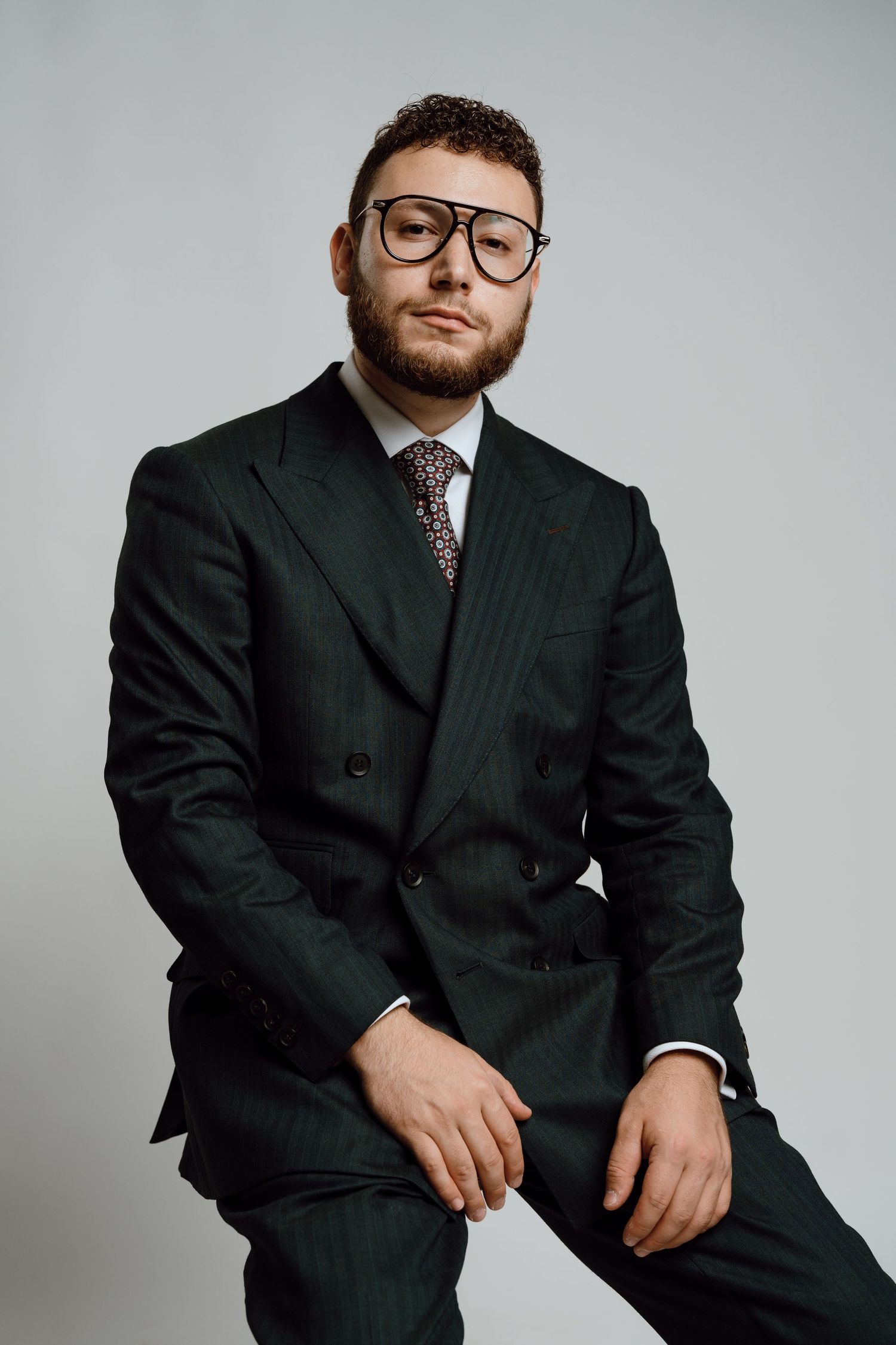 Professional studio portrait of someone in a dark business suit and glasses against a light gray background.