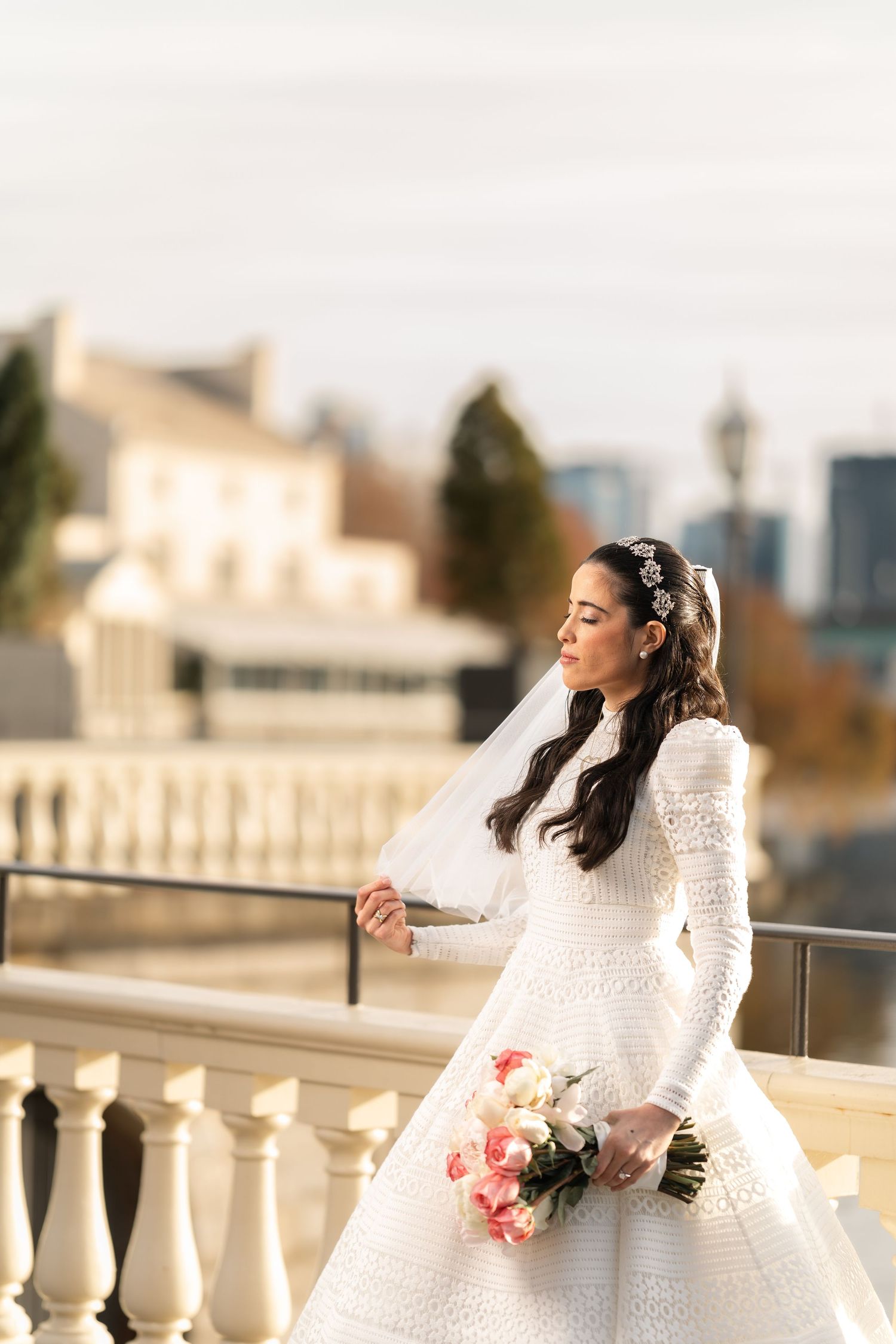 Bridal portrait on a balcony featuring a white lace dress and pink rose bouquet against a city backdrop.