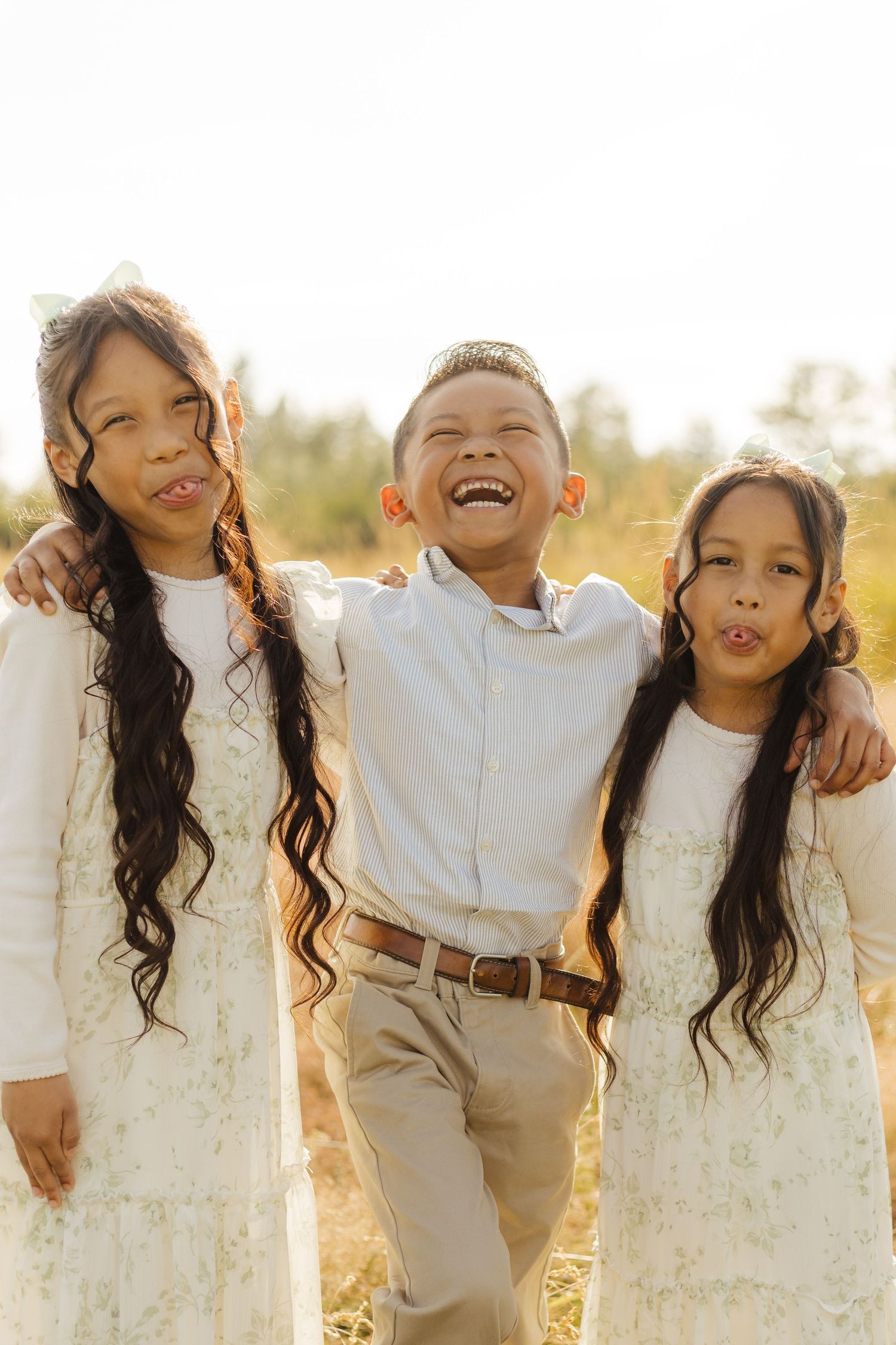 Three siblings in white and khaki outfits making silly faces while posing together in golden sunlight outdoors.