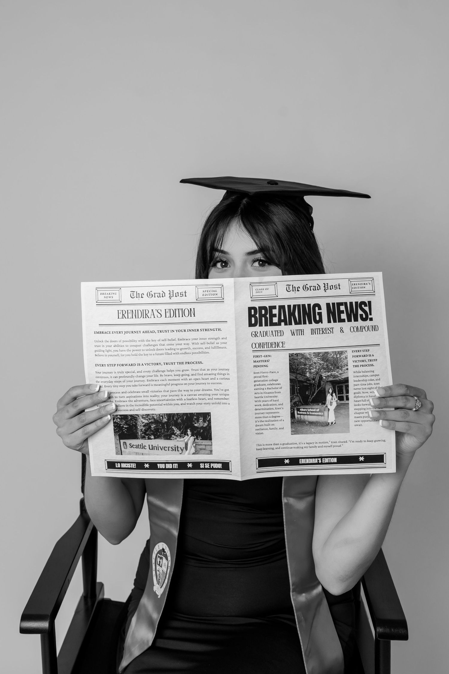 Graduate in cap and gown sitting in chair holding up a Breaking News newspaper in black and white photo.