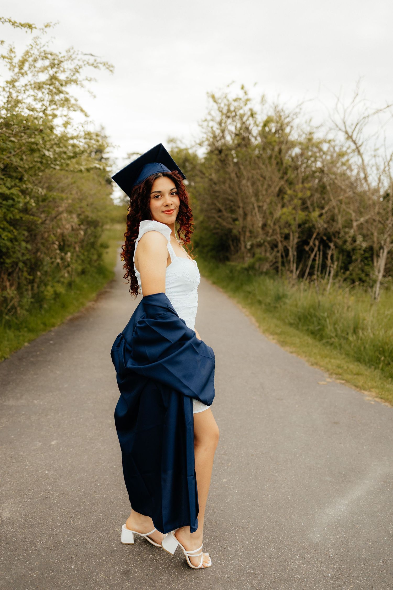 Graduate in a navy cap and gown poses playfully on a rural path surrounded by greenery.