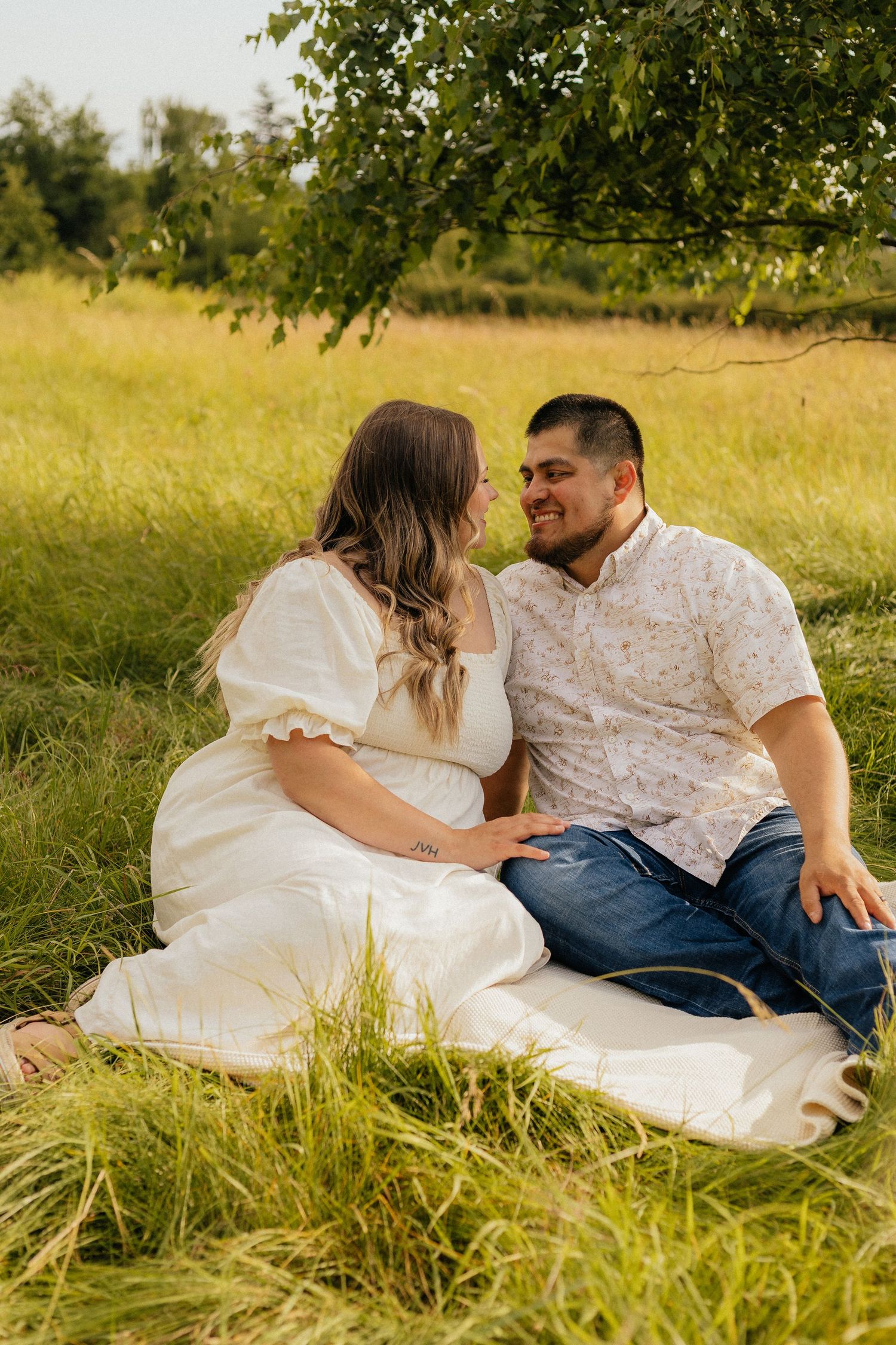 A couple sits together on a white blanket in a sunlit grassy meadow during a romantic outdoor photo session.