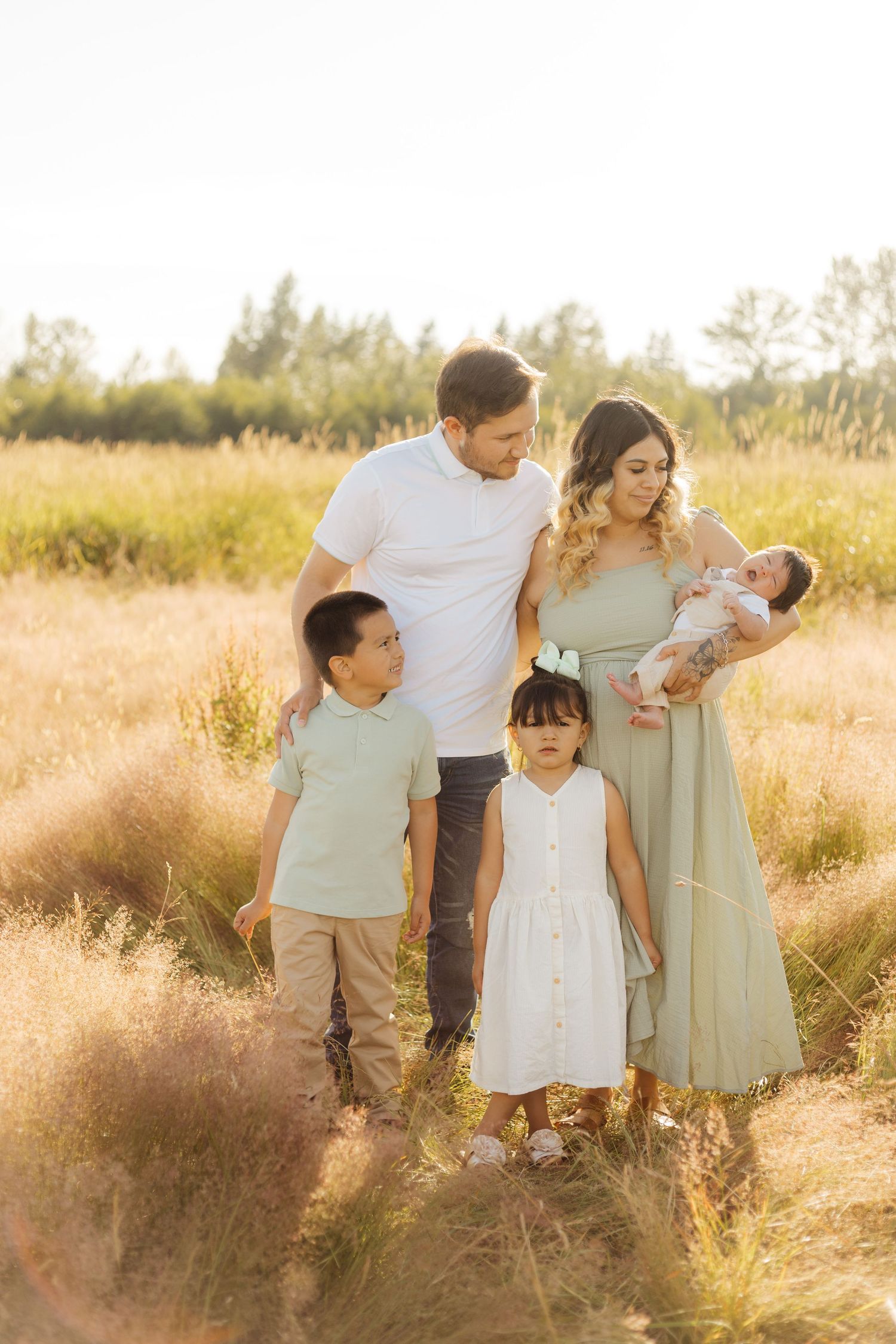 A family wearing white and sage green outfits walks together through a sunlit field of tall golden grass.