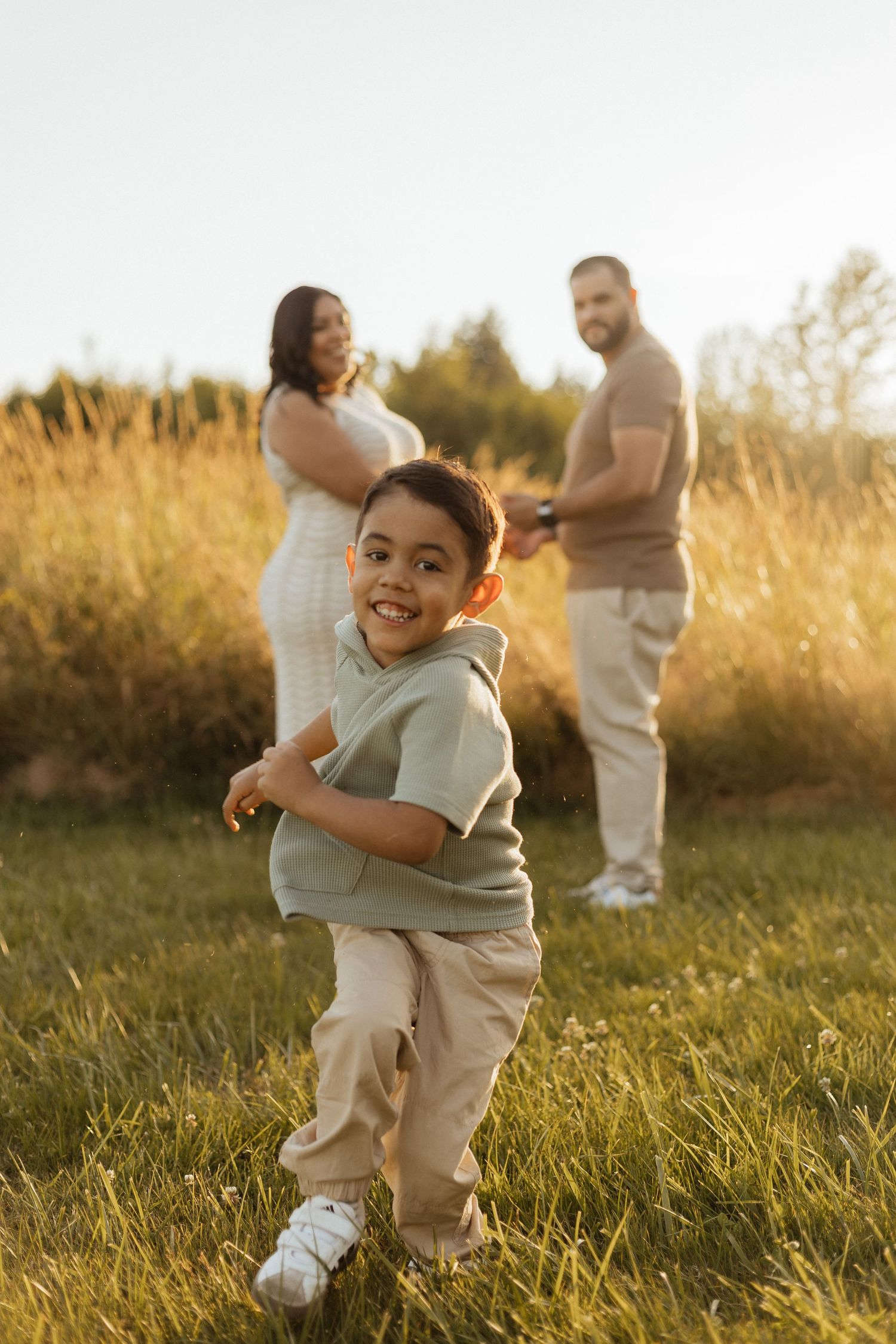 A playful child runs across a sunlit field in a gray hoodie while adults stand in the background during golden hour.