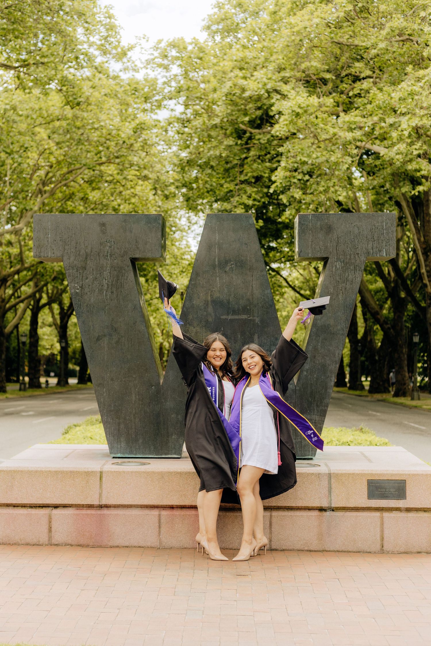 Two graduates celebrate in front of large W statue on University of Washington campus wearing purple graduation stoles.