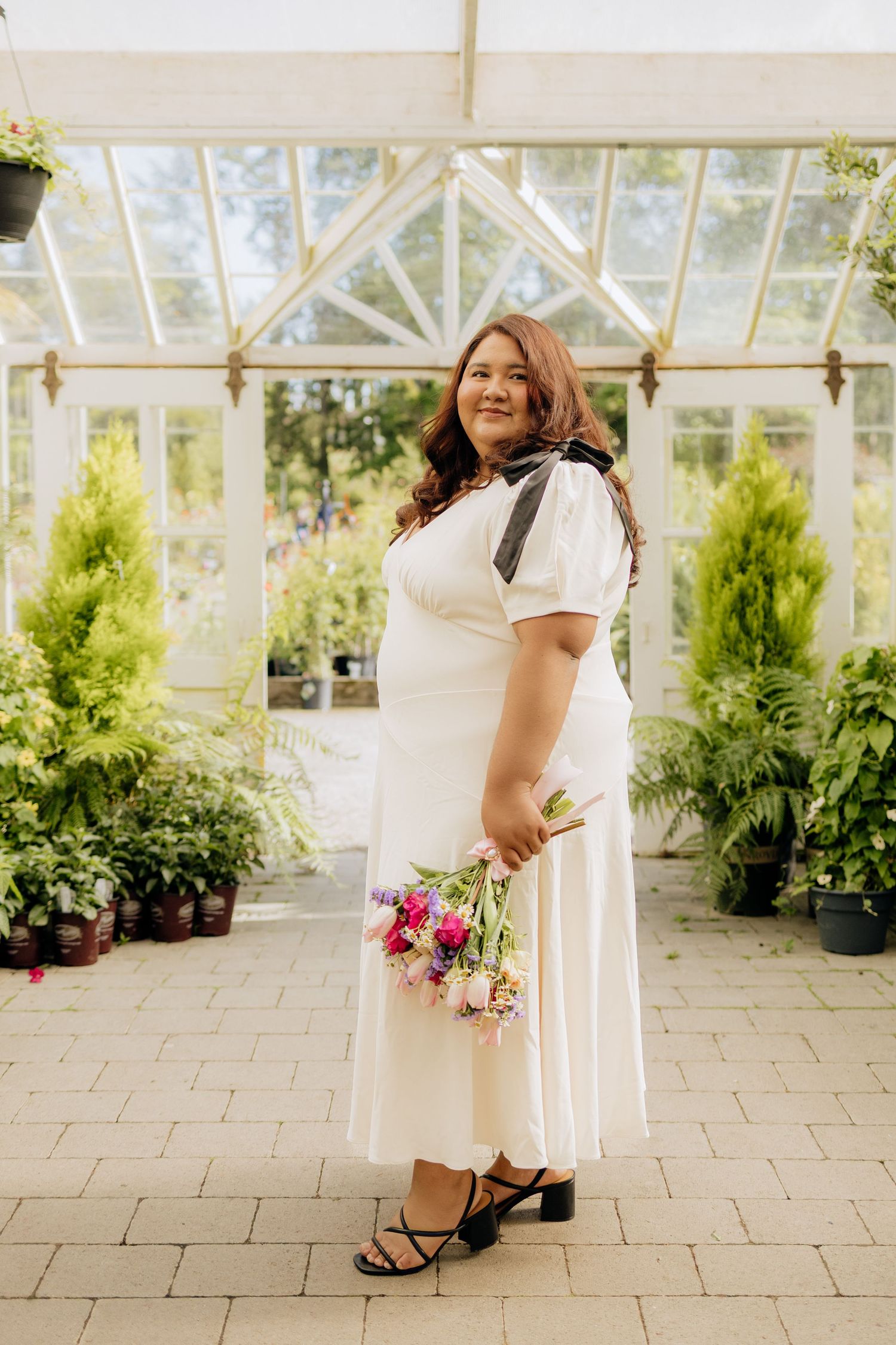 A bridal party in flowing white dresses poses in a sunlit greenhouse with lush greenery and flowers.