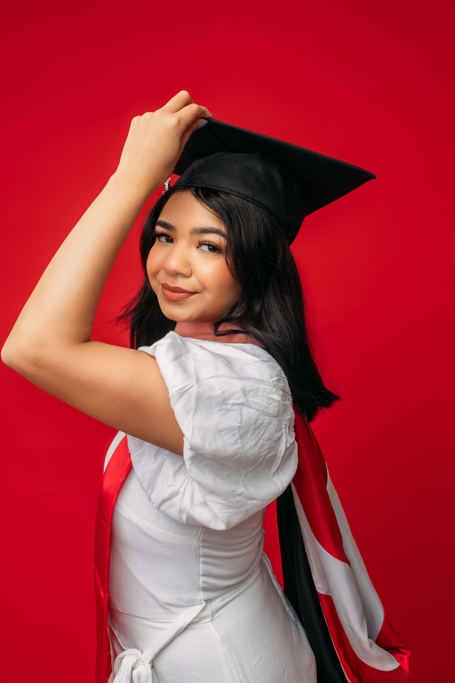 Graduate in white dress poses with graduation cap against vibrant red background.