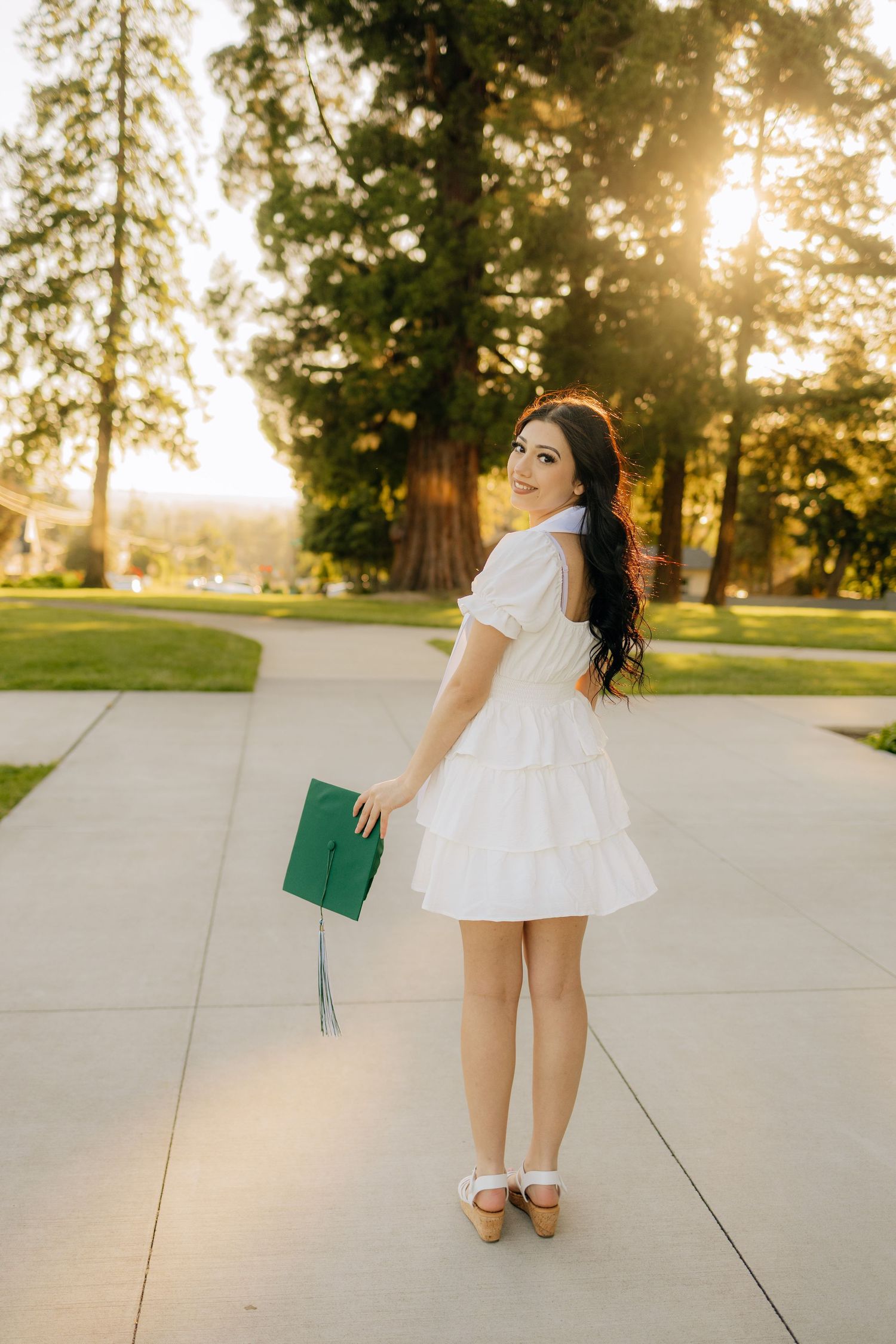 A person in a white sundress holds a green graduation cap while standing on a sunlit campus pathway at golden hour.