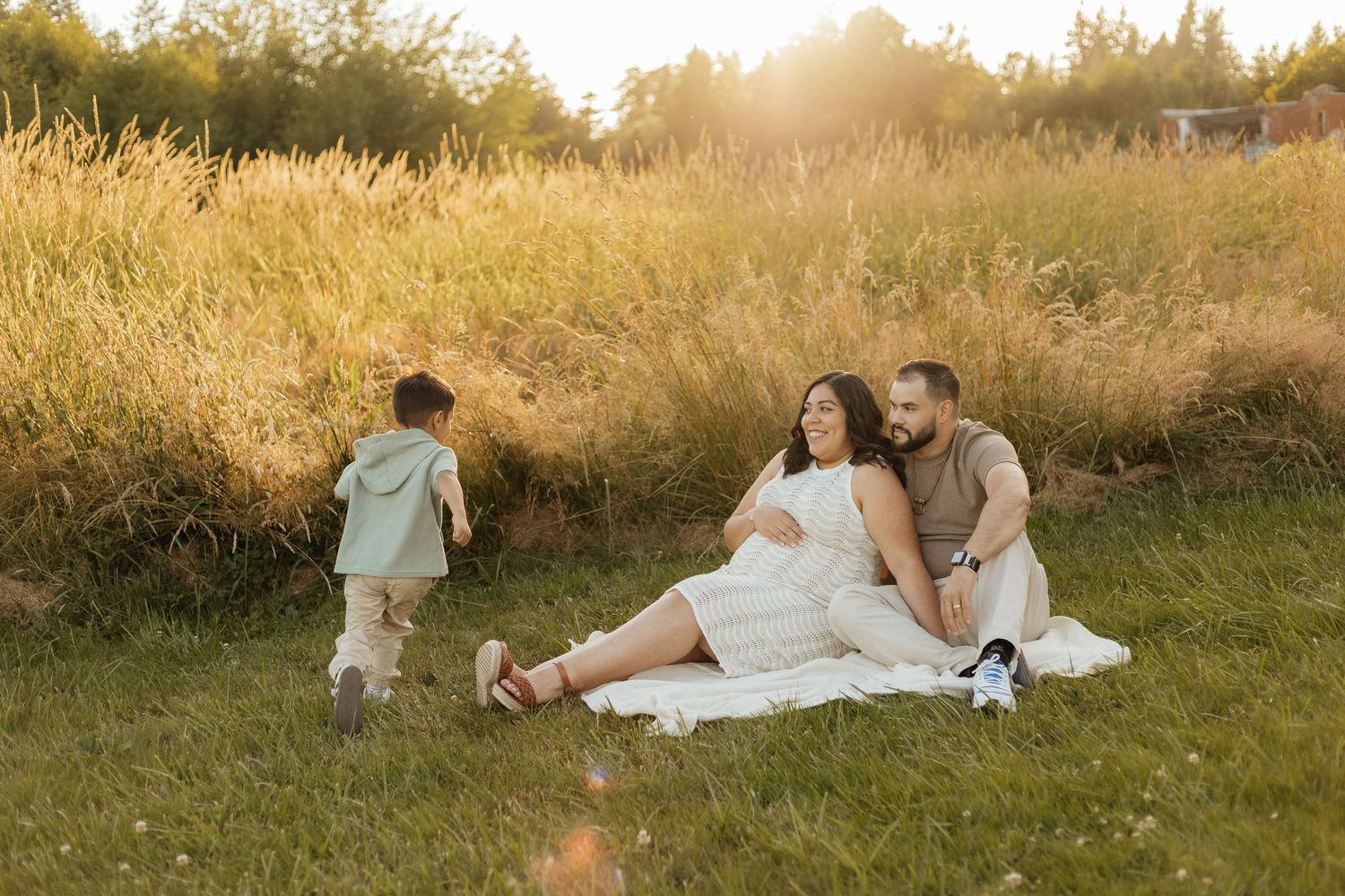 A couple relaxes on a white blanket in a sunlit meadow with tall golden grass at sunset.