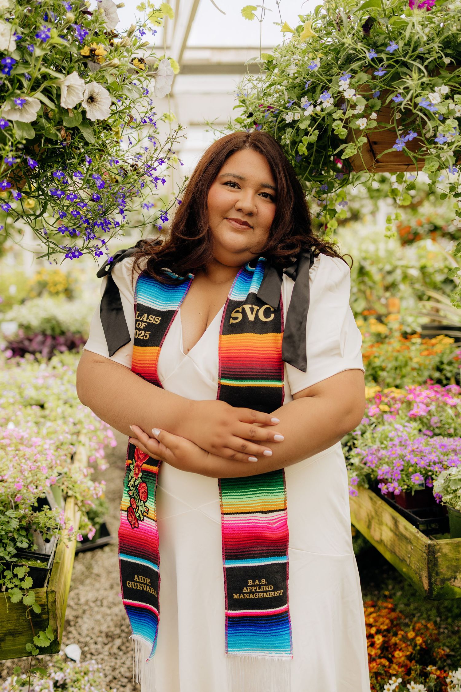 Graduate wearing colorful serape stole and white gown poses amidst vibrant flowers in a greenhouse setting.