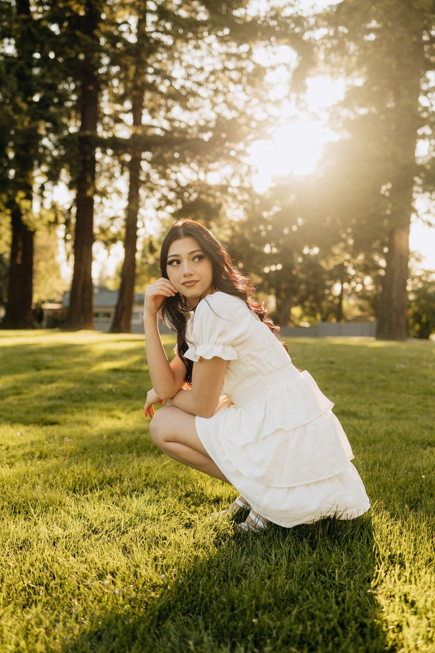 Person in white ruffled dress crouches on sunlit grass with sunlit trees in background during golden hour.