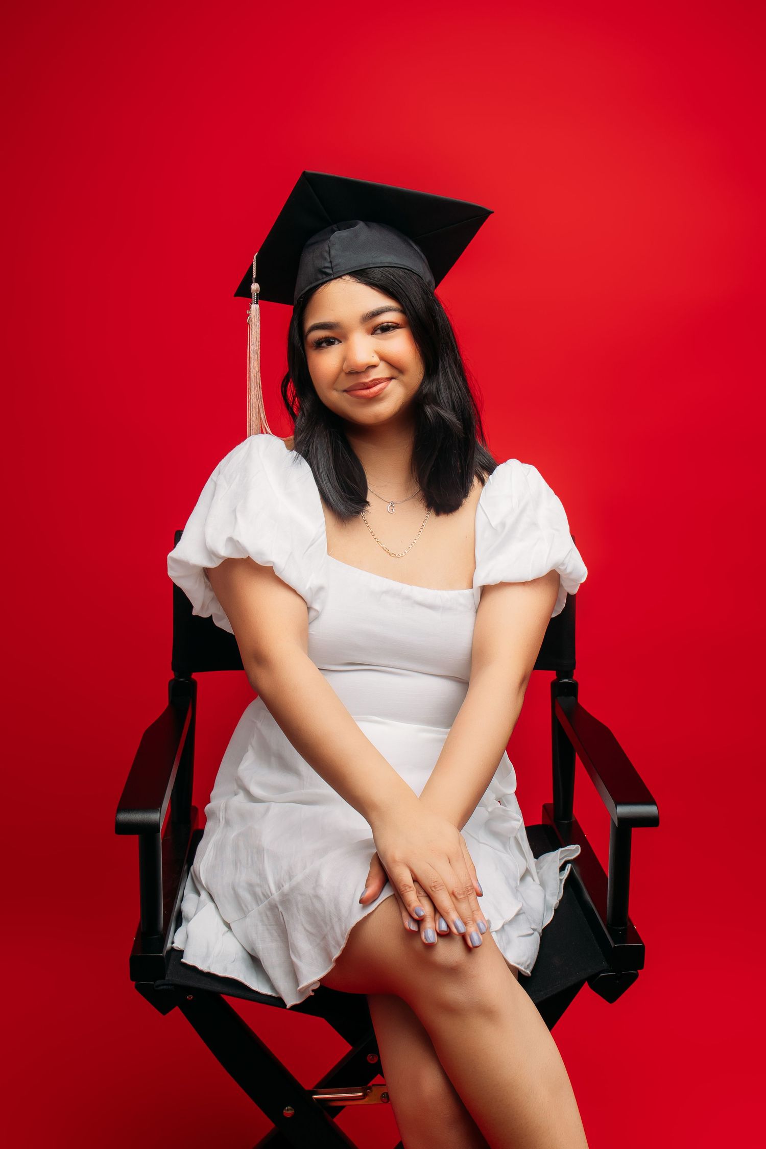 Graduate wearing white dress and mortarboard cap sits in director's chair against vibrant red background.
