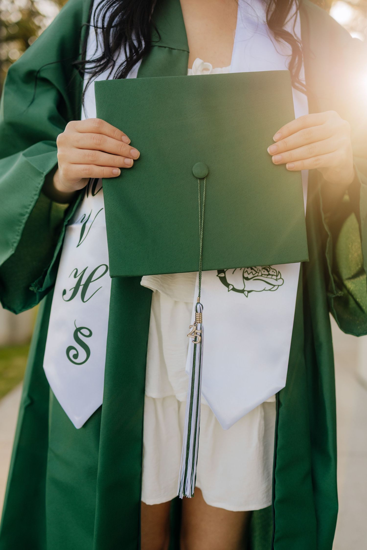 Graduate in green cap and gown holds diploma while wearing a decorated white sash during commencement ceremony.
