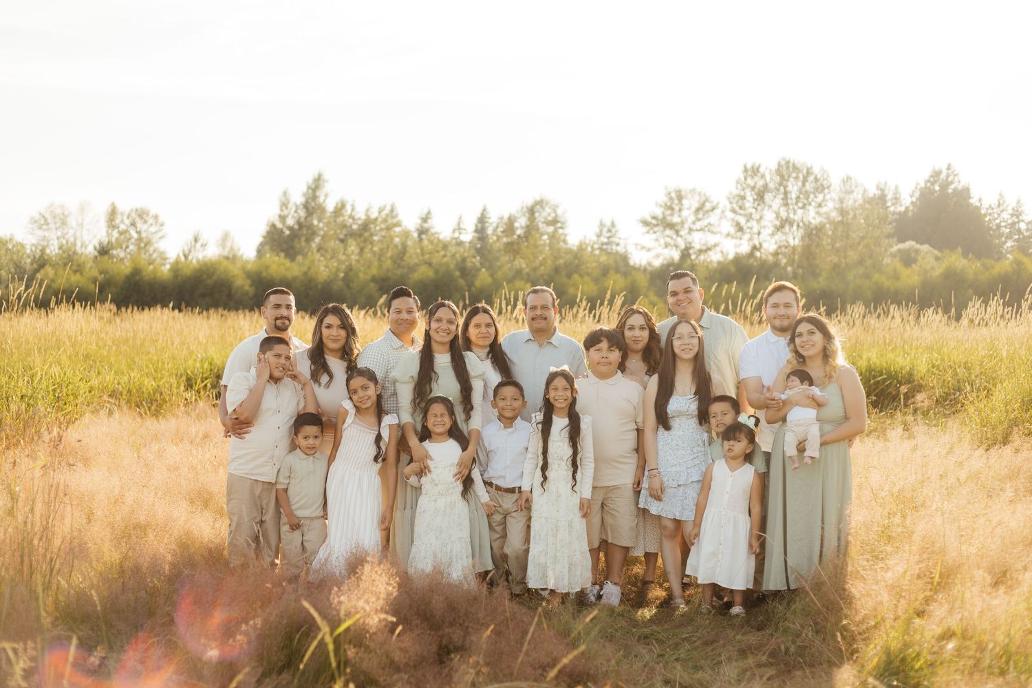 A large group stands together in a sunlit field wearing coordinated white and neutral colored outfits at golden hour.
