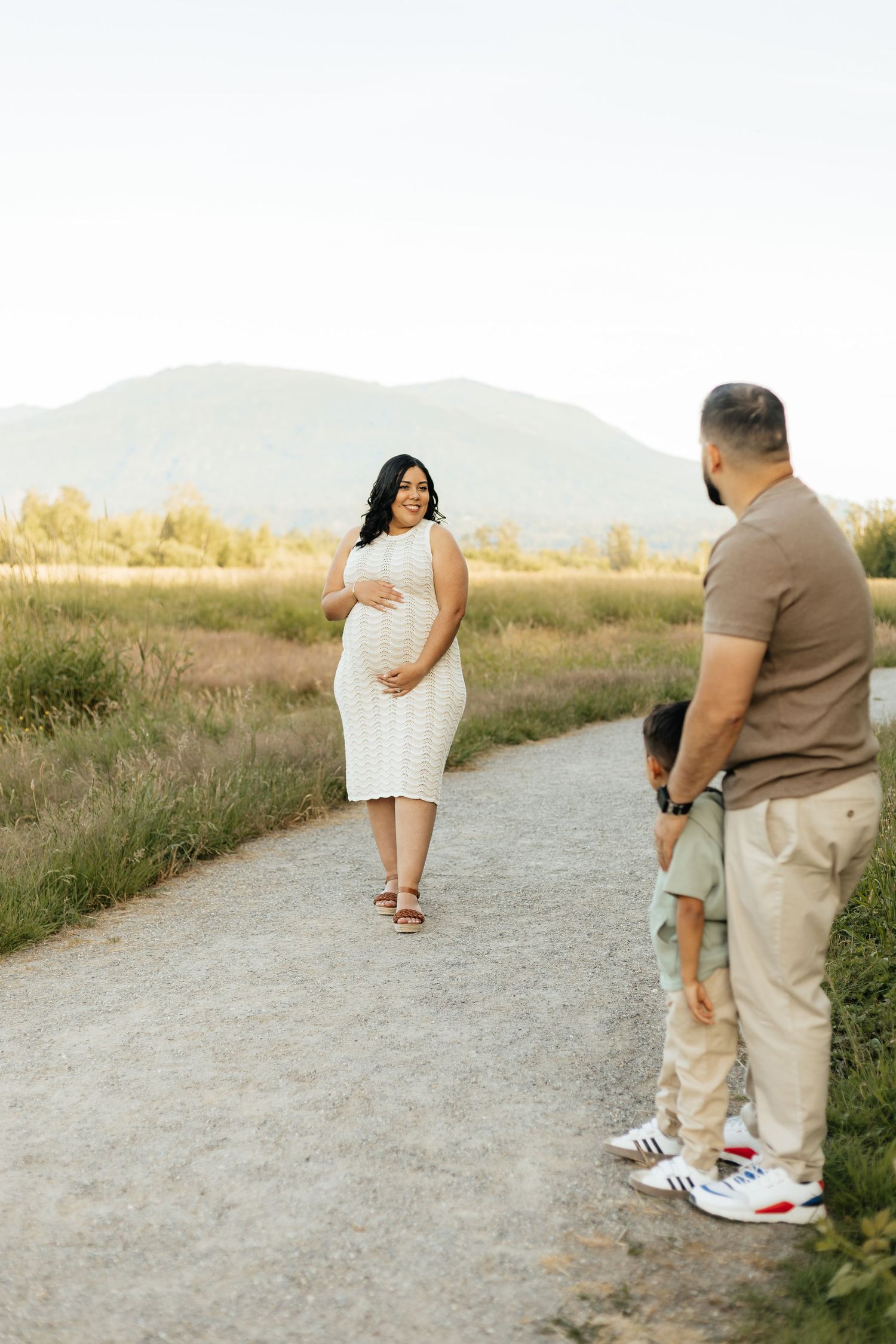 A pregnant woman in a white dress poses on a gravel path while others stand and watch during a sunset photo session.
