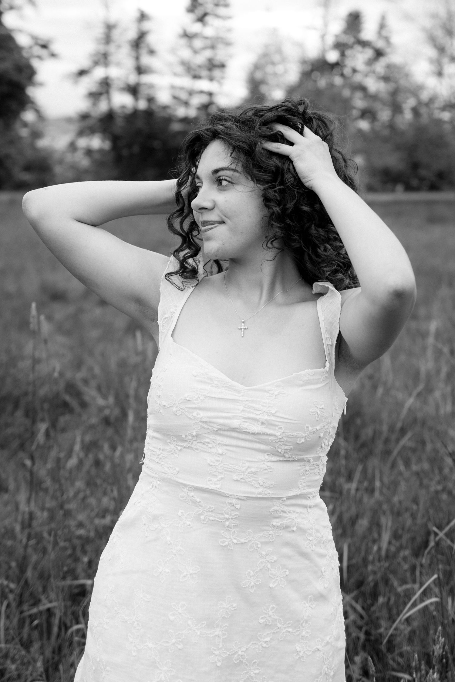 A person with dark curly hair poses in a white sundress outdoors in a field with trees in the background.