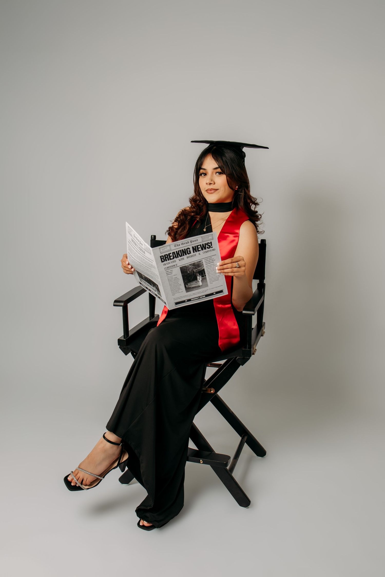 A woman in a red top and black pants poses elegantly with a newspaper in a director's chair against a gray backdrop.
