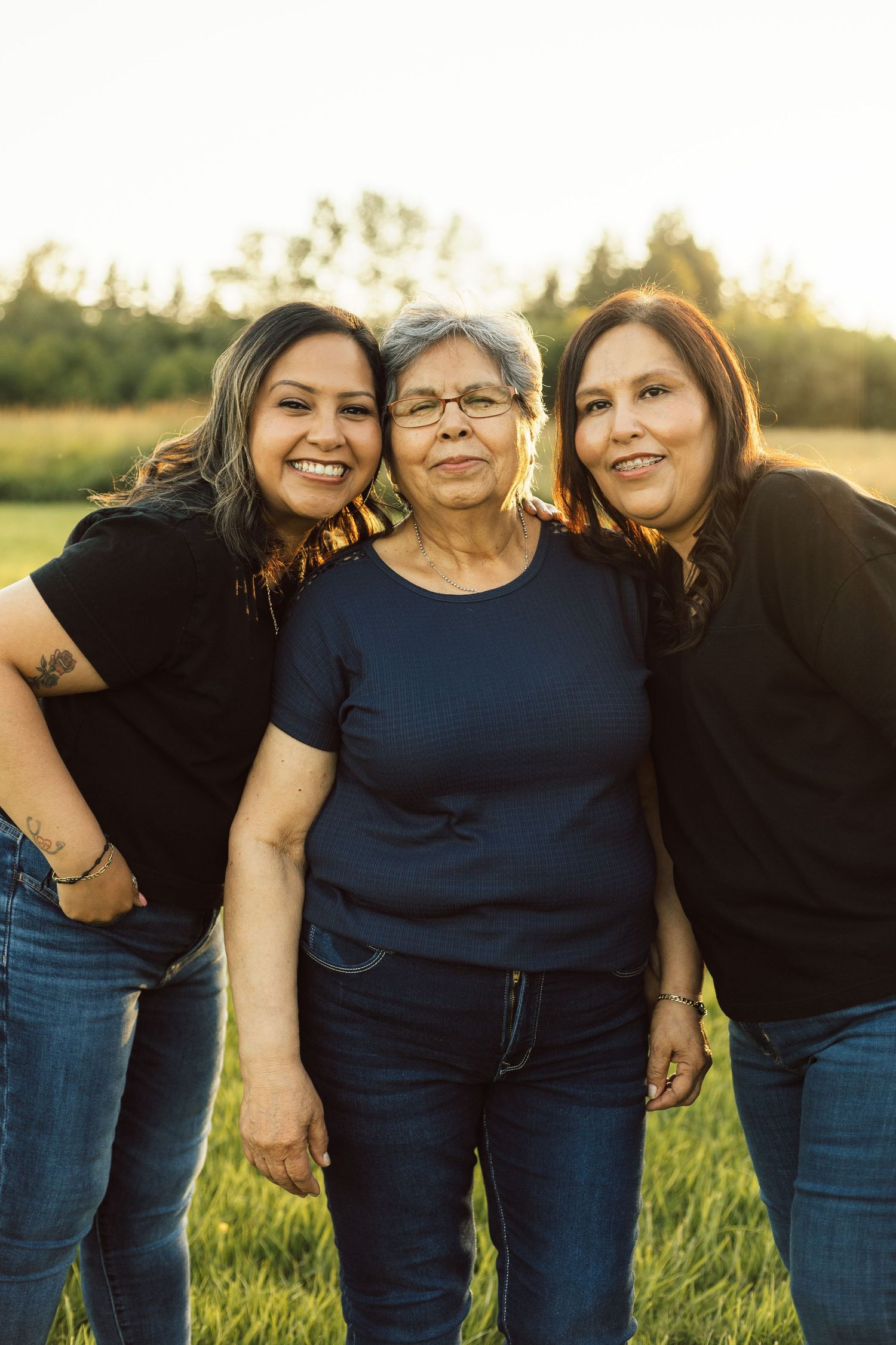 Three generations of women in matching casual outfits pose together in a grassy field at sunset.