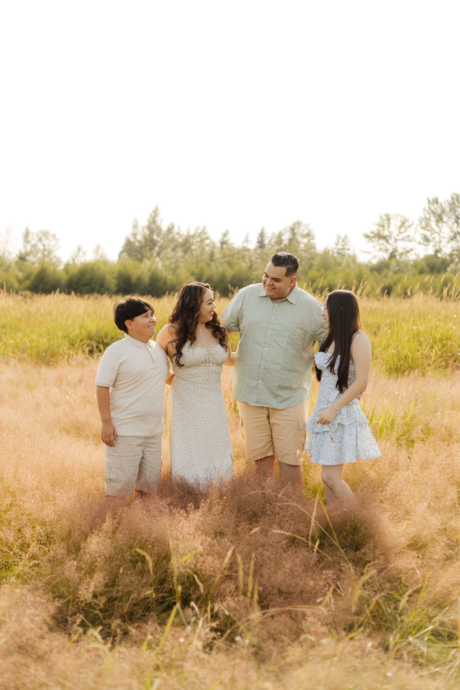 A family stands together in a golden field at sunset surrounded by tall grass and trees in the background.