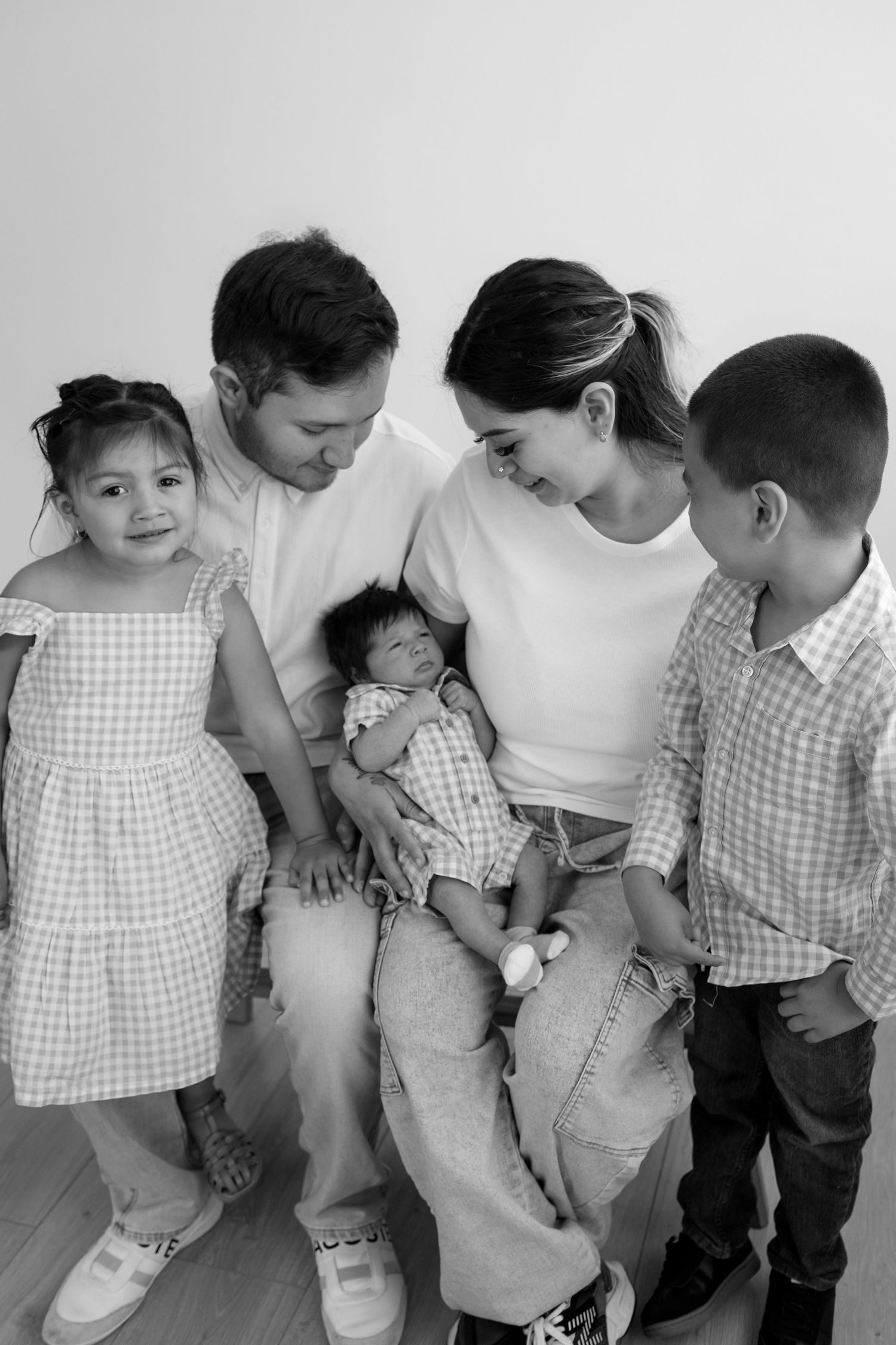Black and white family portrait showing parents looking lovingly at sleeping newborn while siblings gather around.