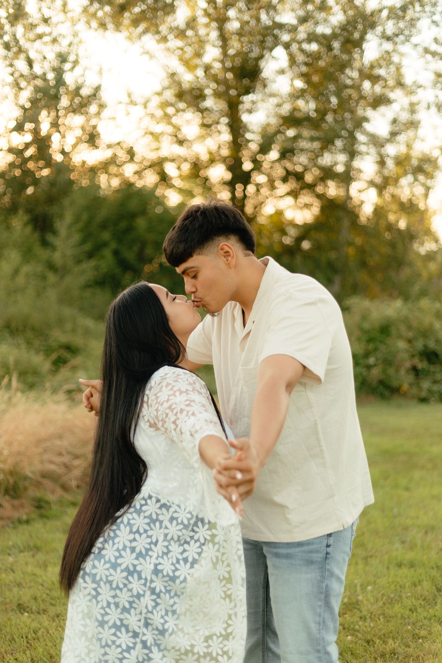 A romantic couple in white outfits share a tender kiss at sunset in a grassy outdoor setting with trees in the background.