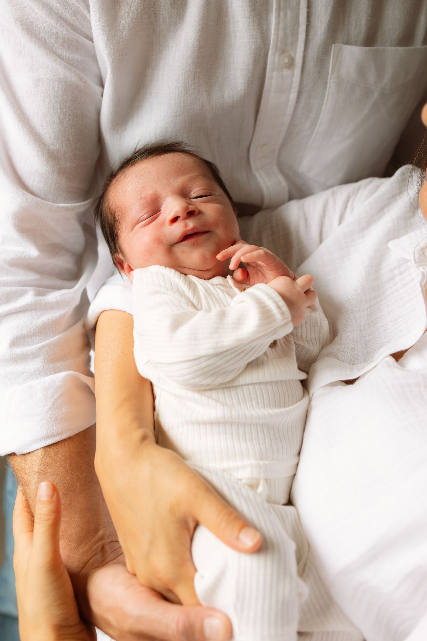 Baby wearing white clothing sleeps peacefully on soft white bedding while being held by adult hands.
