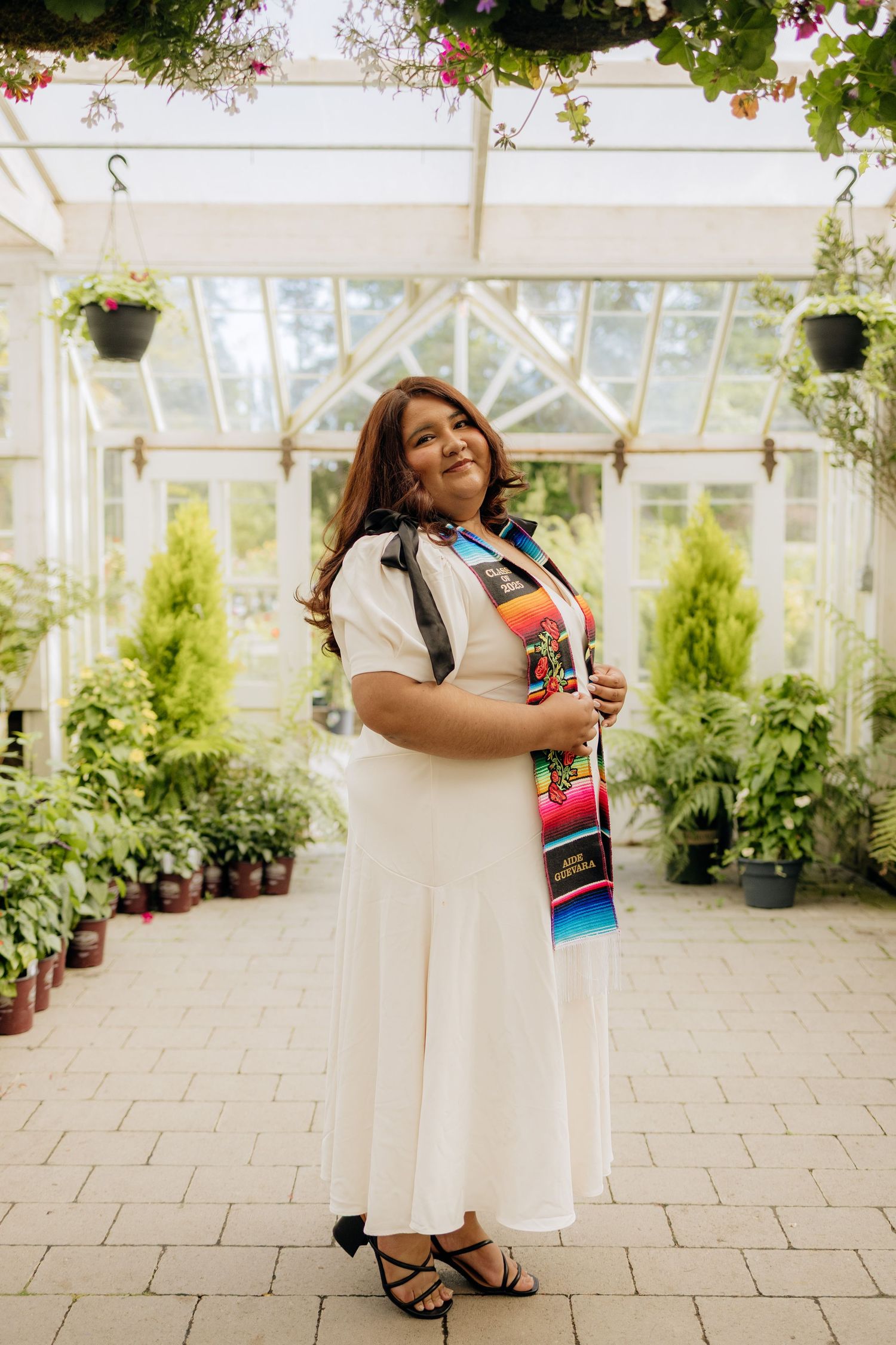 A person in a white dress and colorful sash stands smiling in a greenhouse filled with hanging plants and flowers.