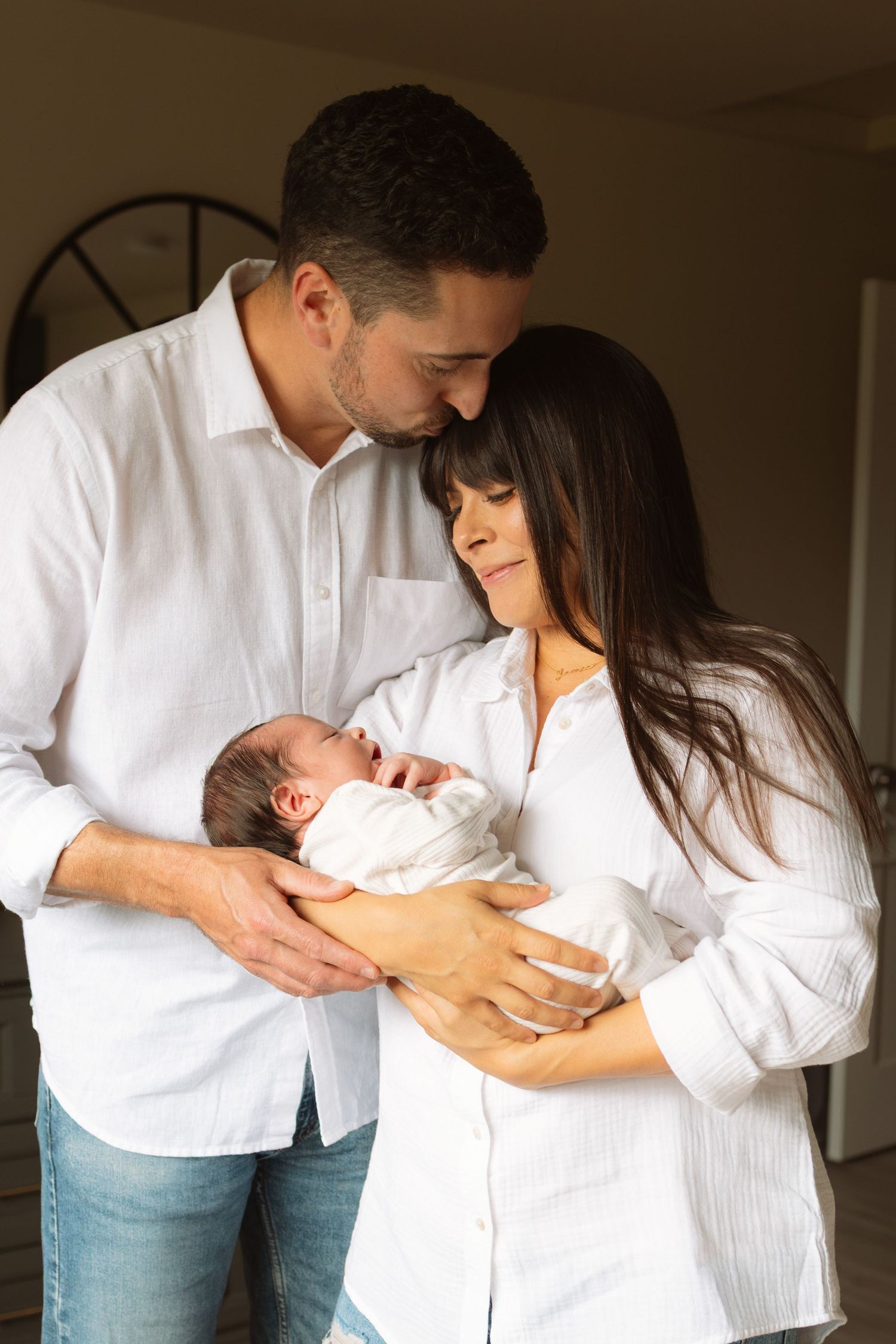 A couple in coordinating white outfits shares tender moments with their newborn baby in a progressive series of intimate poses.