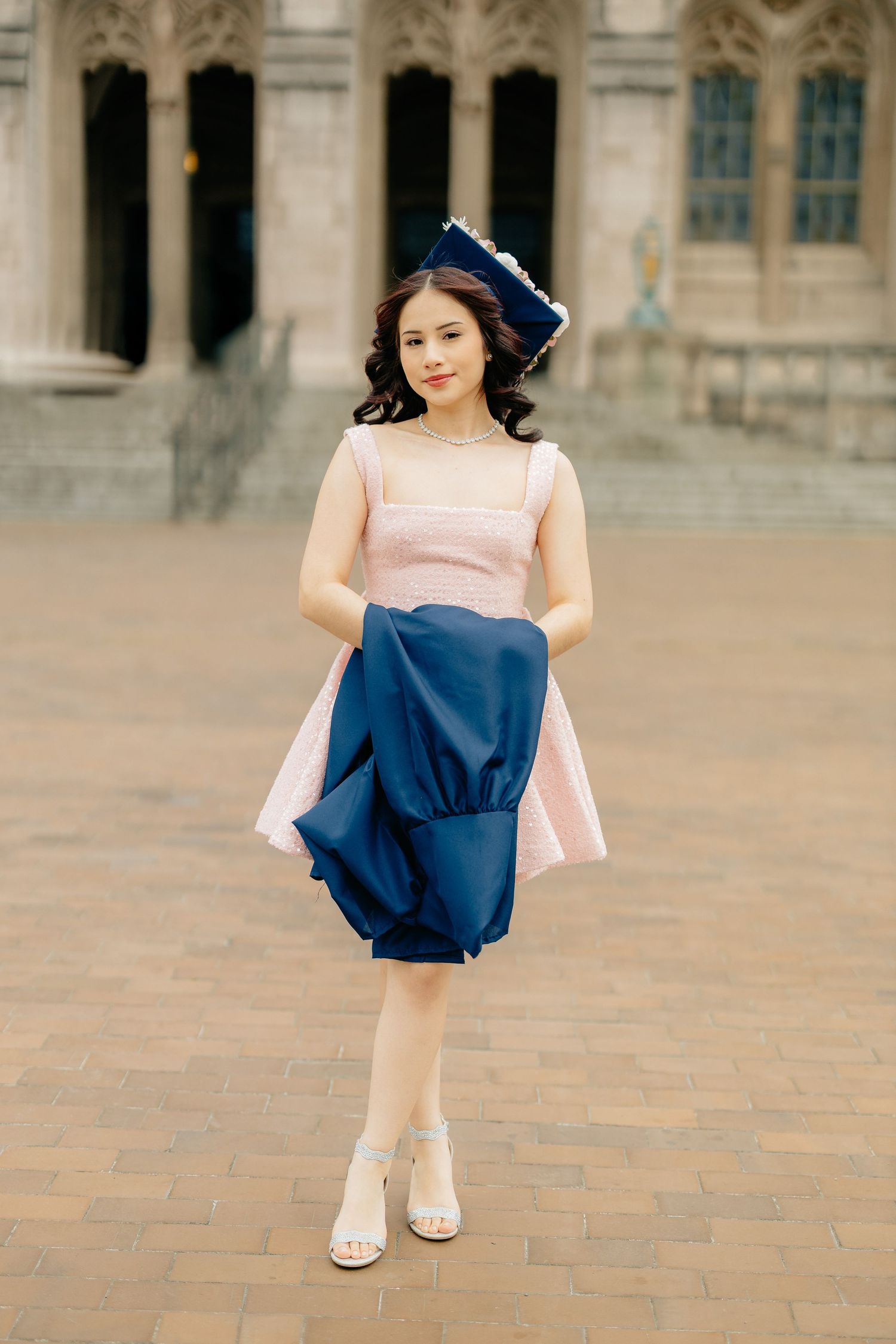 A woman in a pink dress and navy blue accents stands elegantly on brick pavement near a historic building.