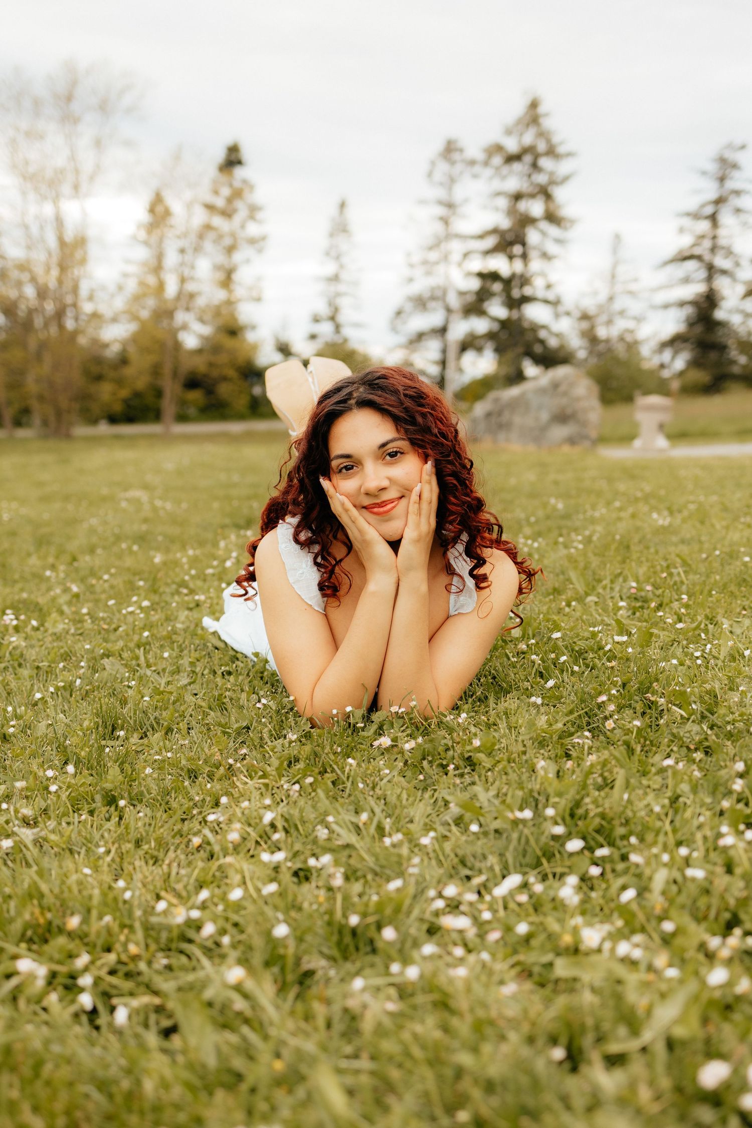 Someone lies in a meadow filled with small white flowers on a sunny spring day, surrounded by tall evergreen trees.
